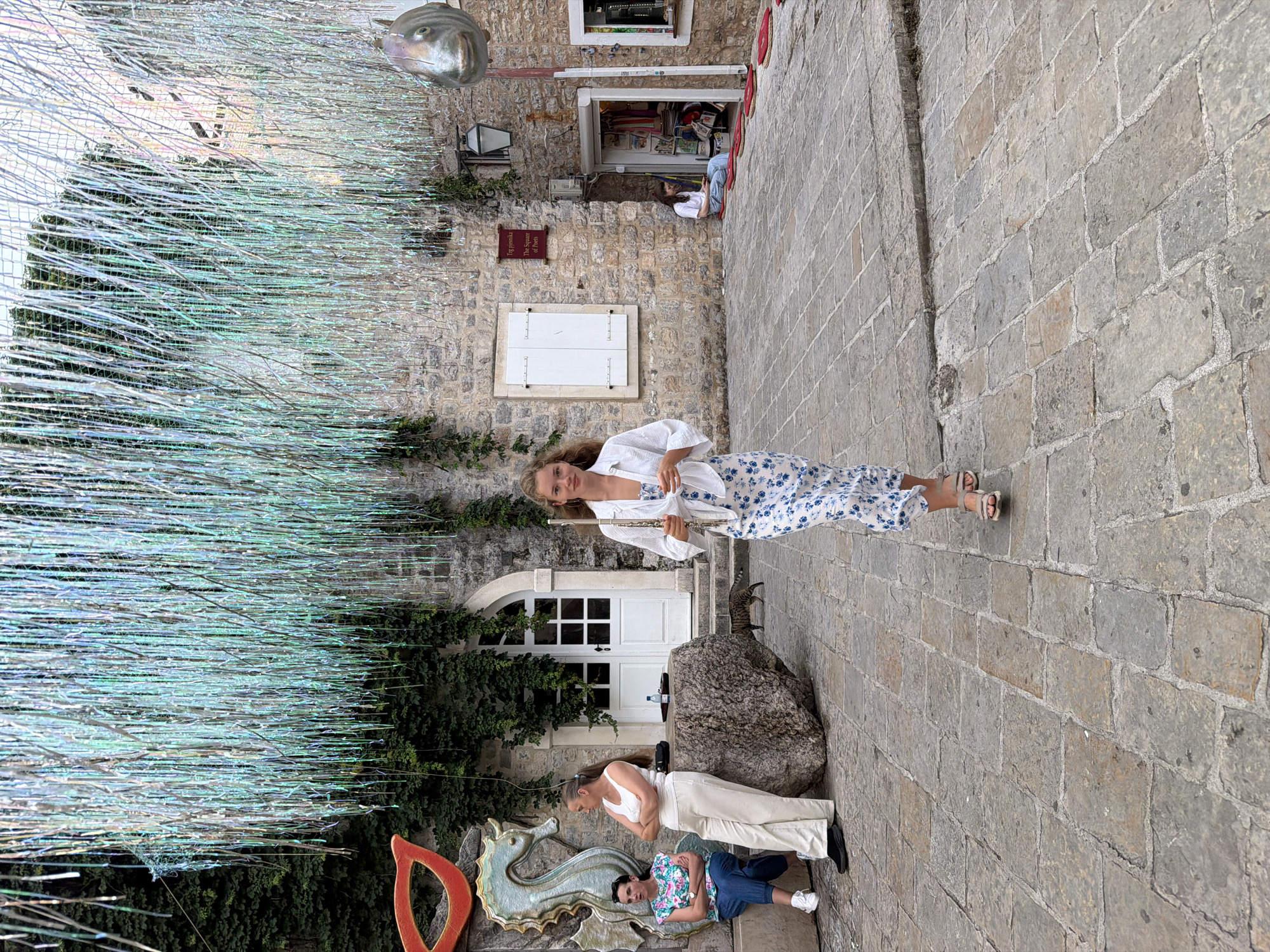 This daytime image captures a vibrant street scene in Budva, Montenegro, likely within its historic old town, characterized by stone buildings and paved pathways. The main subject is a woman with long, curly blonde hair, wearing a white long-sleeved top and a blue and white floral patterned skirt or dress. She stands confidently on the stone pavement, holding a silver flute. She is looking towards the viewer with a slight smile.
Behind her, an old stone building with a white arched door and white shutters is partially covered in dark green ivy. To her right (viewer's left), a large section of iridescent, tinsel-like streamers or decorations hangs overhead, creating a sparkling canopy. High on the right side of the frame, a metallic, fish-shaped sculpture is suspended against the stone wall.
In the lower left portion of the image, two other individuals are present. One woman stands in a white tank top and light-colored pants, while another person sits on the ground in a floral shirt and blue shorts. They are positioned near a large natural rock and a colorful, sculpted seahorse decoration. Partially visible next to the seahorse is an orange letter "D" sculpture.
Further in the background, another stone building features a small, open shop or kiosk, with merchandise visible inside and red mats laid out on the pavement. A small cat is also visible near the white door behind the main woman. A dark sign on the stone wall reads "THE SQUARE of Poets." The ground throughout the scene is paved with rough-hewn stone blocks. The overall atmosphere is relaxed and picturesque.