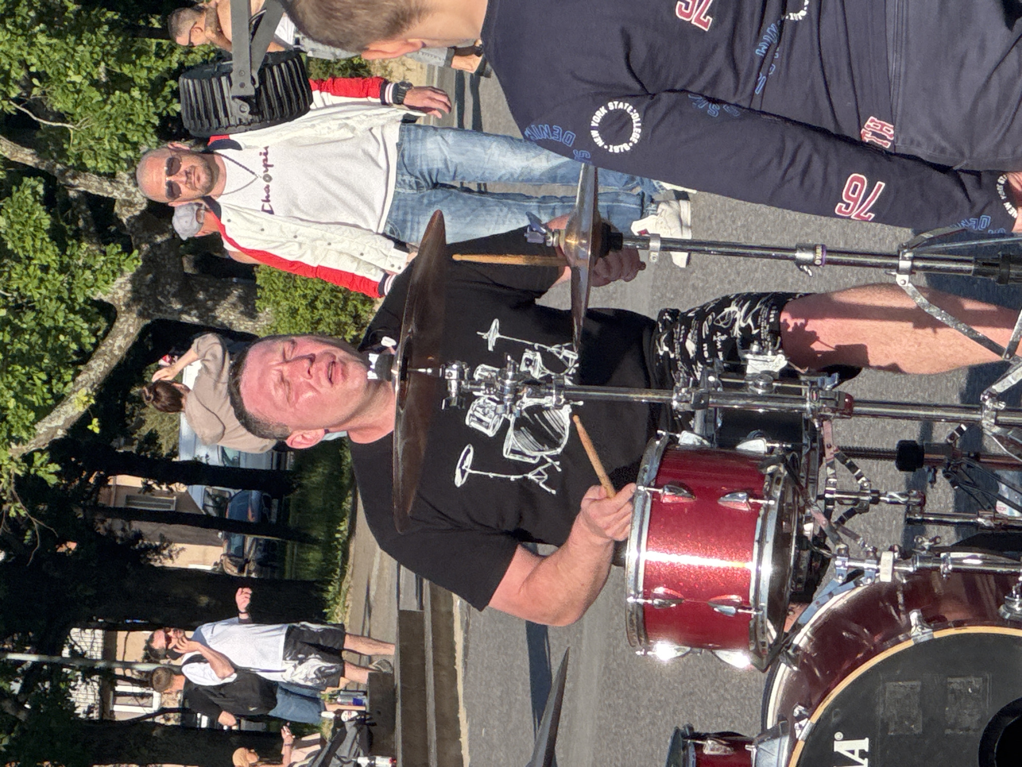 A man wearing a black t-shirt with a drum set graphic is playing a drum kit outdoors. He is sweating and has his eyes closed. He is wearing patterned shorts and his legs are visible. He is holding drumsticks and hitting a red drum. The drum kit is metallic and shiny. Other people are visible in the background, some blurred. One man in the background is wearing a white jacket with red stripes and jeans. Another man in the background is wearing a white t-shirt and shorts and is on the phone. The setting appears to be a public area with trees and a street. The image appears to be taken during daylight with sunlight casting shadows. There is no visible indication of the specific location in Tallinn, Estonia.