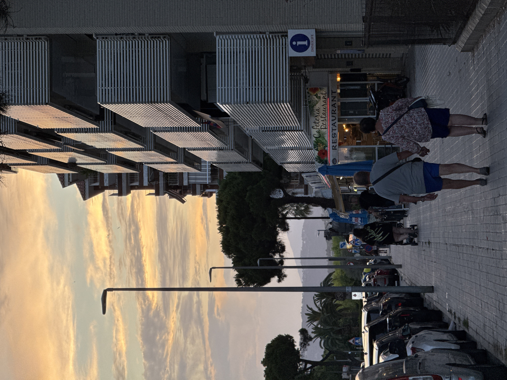 This is a medium shot of a street scene in Blanes, Spain, at sunset. The sky is filled with pale orange and yellow clouds, with several distant birds visible. On the right side of the image is a multi-story building with numerous balconies, some of which have white slatted railings. Below the balconies, there is a storefront with a sign that reads "Cafeteria Restaurant" and "RESTAURANT." A small blue umbrella is visible in front of the establishment. To the left of the building, a sidewalk runs parallel to the street. A man and a woman are walking hand-in-hand on the sidewalk, facing away from the camera. The man is wearing a grey t-shirt and blue shorts, and the woman is wearing a floral print shirt and blue shorts. A woman with a baby stroller is walking ahead of them, also facing away from the camera. A line of parked cars is visible on the left side of the sidewalk, with palm trees and other foliage in the background. In the distance, mountains are silhouetted against the twilight sky. A lamppost stands on the sidewalk near the center of the image.