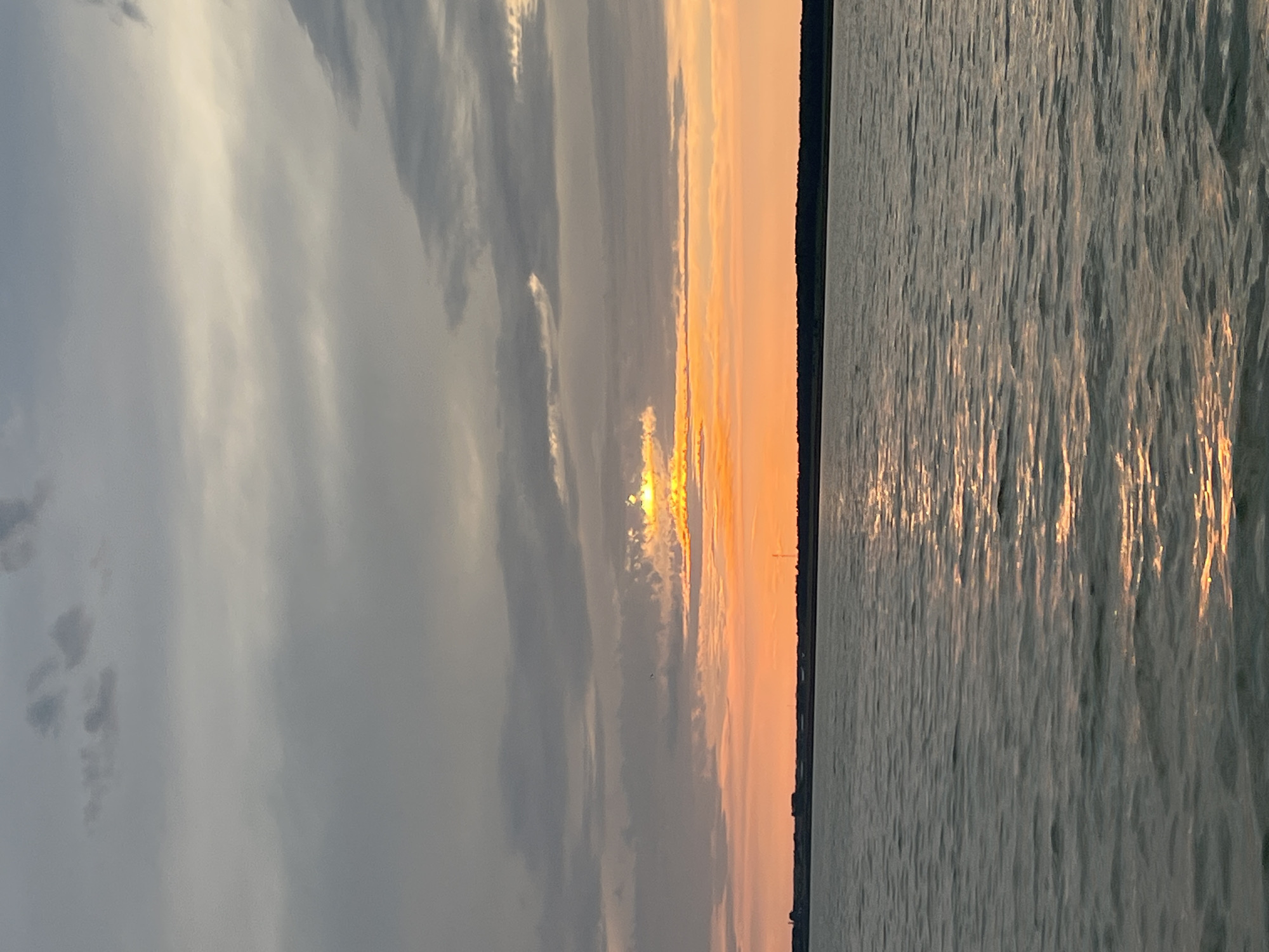 The image shows a body of water with a shoreline in the distance under a cloudy sunset sky. The sun is low on the horizon, casting a warm orange glow through breaks in the clouds, creating streaks of light on the water's surface. The water is choppy, reflecting the colors of the sky. The shoreline appears to be a strip of dark vegetation with some structures visible in the distance. The overall scene evokes a sense of tranquility and the end of the day. There are no people or animals visible in the image. The location is not identifiable as Borovoe, Russia.
