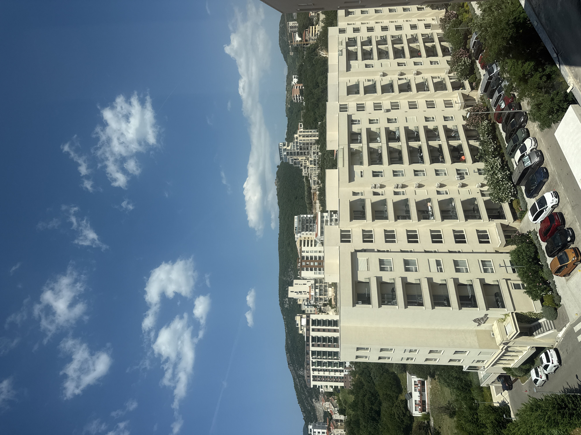 A high-angle, medium shot captures a segment of the city of Boreti, Montenegro, under a bright, partly cloudy sky. The foreground is dominated by a large, cream-colored apartment building with numerous balconies. Below the building, a parking lot is filled with various vehicles, including white SUVs, a dark blue sedan, a silver van, a dark gray SUV, a black car, a red car, a white car, and a small orange car. Several trees and flowering bushes are visible surrounding the parking area. In the mid-ground, more apartment buildings of varying heights and architectural styles are nestled into a lush green hillside. The background features rolling, tree-covered hills under a blue sky scattered with white, fluffy clouds. One building in the mid-ground is particularly distinctive with its layered, stepped design. The overall scene depicts a residential area within a hilly, coastal environment.