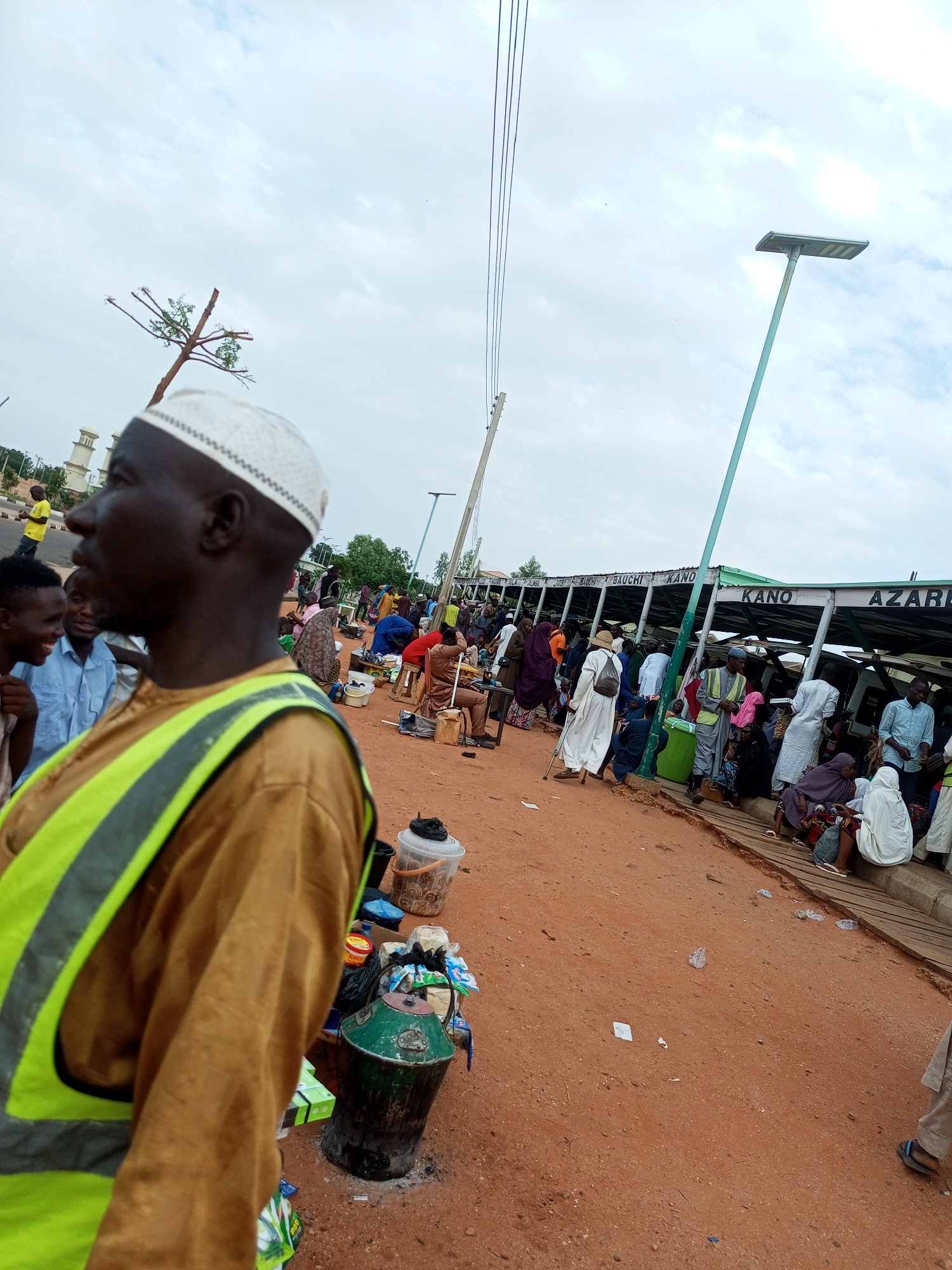 The image depicts an outdoor scene with a crowd of people and various objects, likely in Gombe, Nigeria, given the context of the prompt mentioning the city. In the foreground, a man wearing a white kufi and a high-visibility yellow vest over a brown garment is partially visible, facing left. Behind him, a group of people are gathered, some appearing to be in conversation.
Further into the background, a covered structure with signage indicating destinations such as "KANO" and "AZARE" suggests a bus station or market area. Numerous individuals, many dressed in traditional attire, are seated or standing around this structure. Some appear to be waiting, while others may be engaged in commerce or social interaction. Various items are scattered on the ground, including buckets, containers, bags, and what appear to be food items or goods for sale. The ground is predominantly reddish-brown earth. In the distance, a few trees and what looks like a building or tower are visible. The sky is overcast with clouds. The overall atmosphere suggests a bustling public space, possibly a transportation hub or a marketplace.