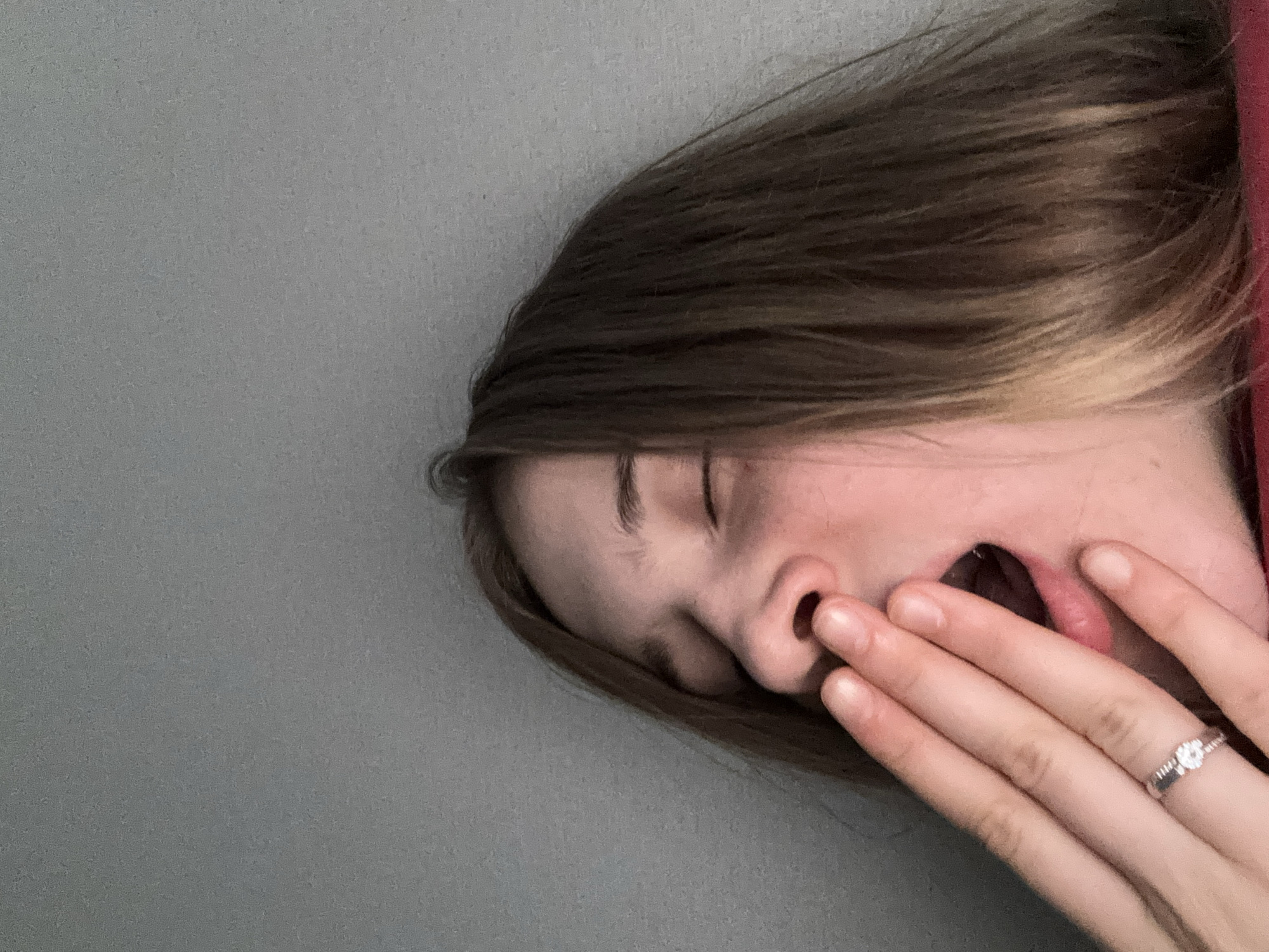 The image shows a close-up of a person's face, with their eyes closed and their mouth open in a yawn. Their hand is covering their mouth, and a ring is visible on one of their fingers. The background is a plain gray wall. The person appears to be indoors. There is no indication of the city of Vsevolozhsk, Russia in the image.