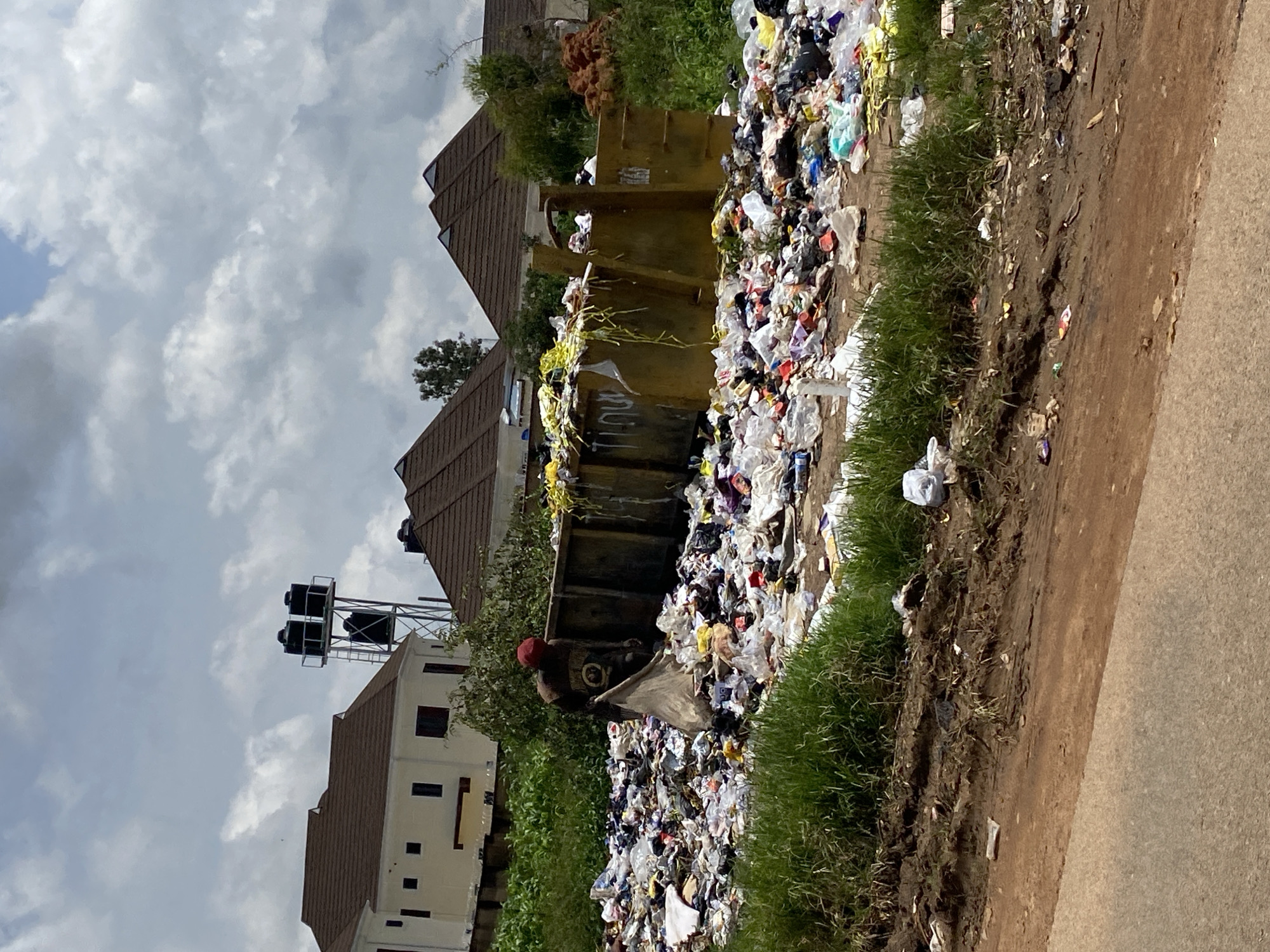 In Gura Top, Nigeria, a scene of heavy refuse accumulation is depicted, with a large yellow skip overflowing with garbage. A lone individual, wearing a red cap and dark clothing, is observed amongst the debris, holding a large plastic bag, possibly engaged in sorting or collecting refuse. The surrounding environment shows signs of neglect, with a substantial amount of trash scattered on the ground and encroaching on a grassy verge. In the background, modern residential buildings with brown tiled roofs stand in contrast to the unsanitary conditions in the foreground, and a water tower with multiple black tanks is visible. The sky is overcast with heavy clouds.
