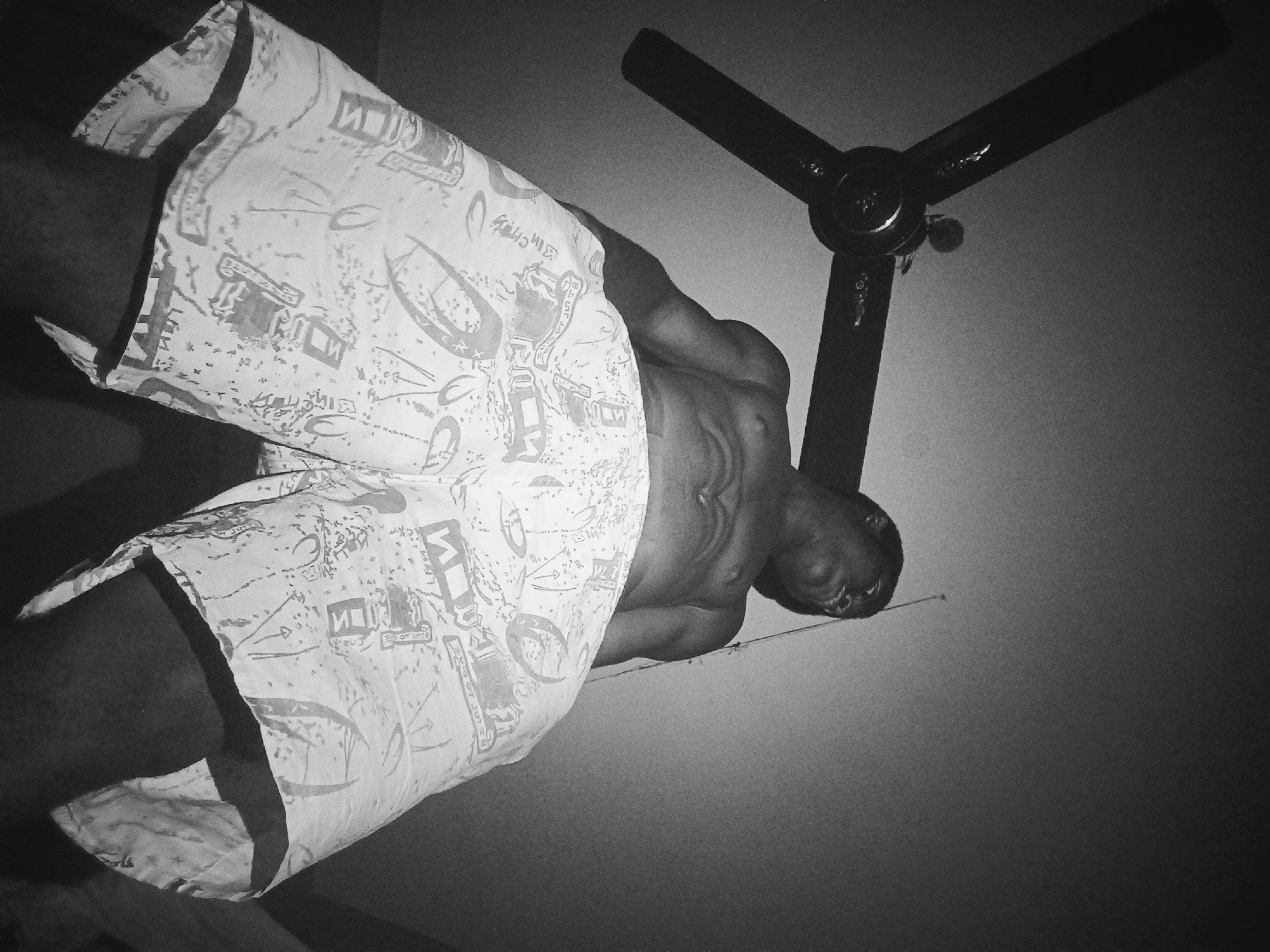 A low-angle, black and white photo shows a partially clothed dark-skinned person standing in a room in Nkpor, Nigeria. The person is wearing patterned shorts and their chest and abdomen are visible. Above the person, a ceiling fan with three blades is mounted. The background consists of a plain wall.