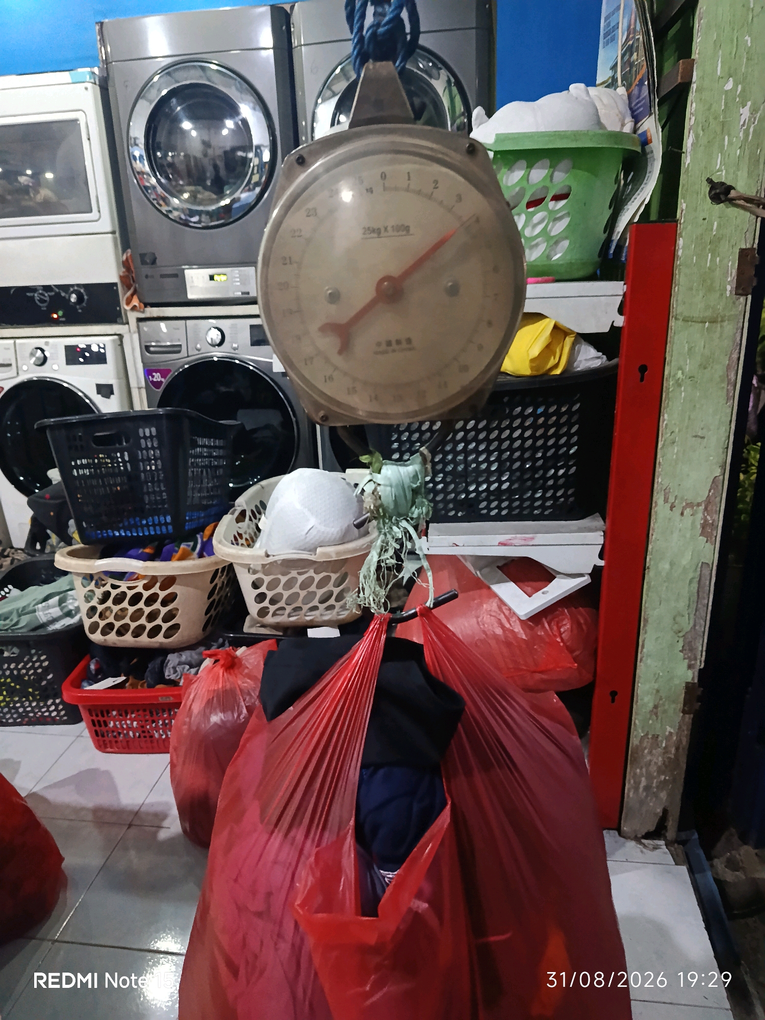 The image depicts a laundry area, likely a commercial laundry service, in Keboansikep, Indonesia. The scene is cluttered with laundry baskets, large red plastic bags filled with clothes, and several washing machines stacked in the background. A mechanical scale with a dial, reading approximately 25 kg, is prominently displayed in the foreground, suggesting a weighing station. Various laundry baskets, some empty and some containing clothes or white items, are scattered throughout. A green plastic laundry basket with a white item inside is visible on a shelf. The floor is tiled. The overall impression is one of a busy, functional space dedicated to handling laundry.