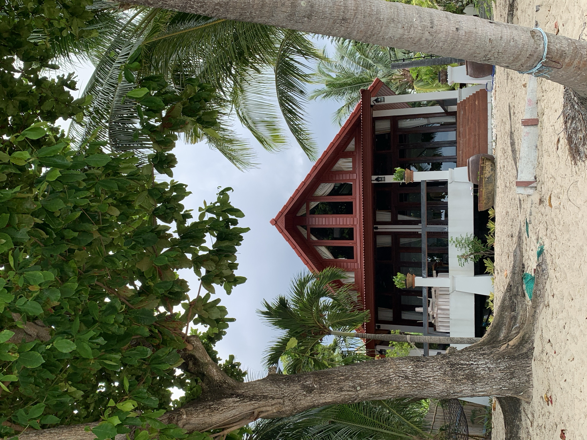 The image depicts a two-story structure with a red gable roof, dark-framed windows, and white support columns. Plants are visible on the balcony. The building is framed by tropical trees, including palm trees and trees with broad green leaves. The scene suggests a location in Baan Tai, Thailand, likely near a beach, as evidenced by the sandy ground visible near the trees. No people are visible.