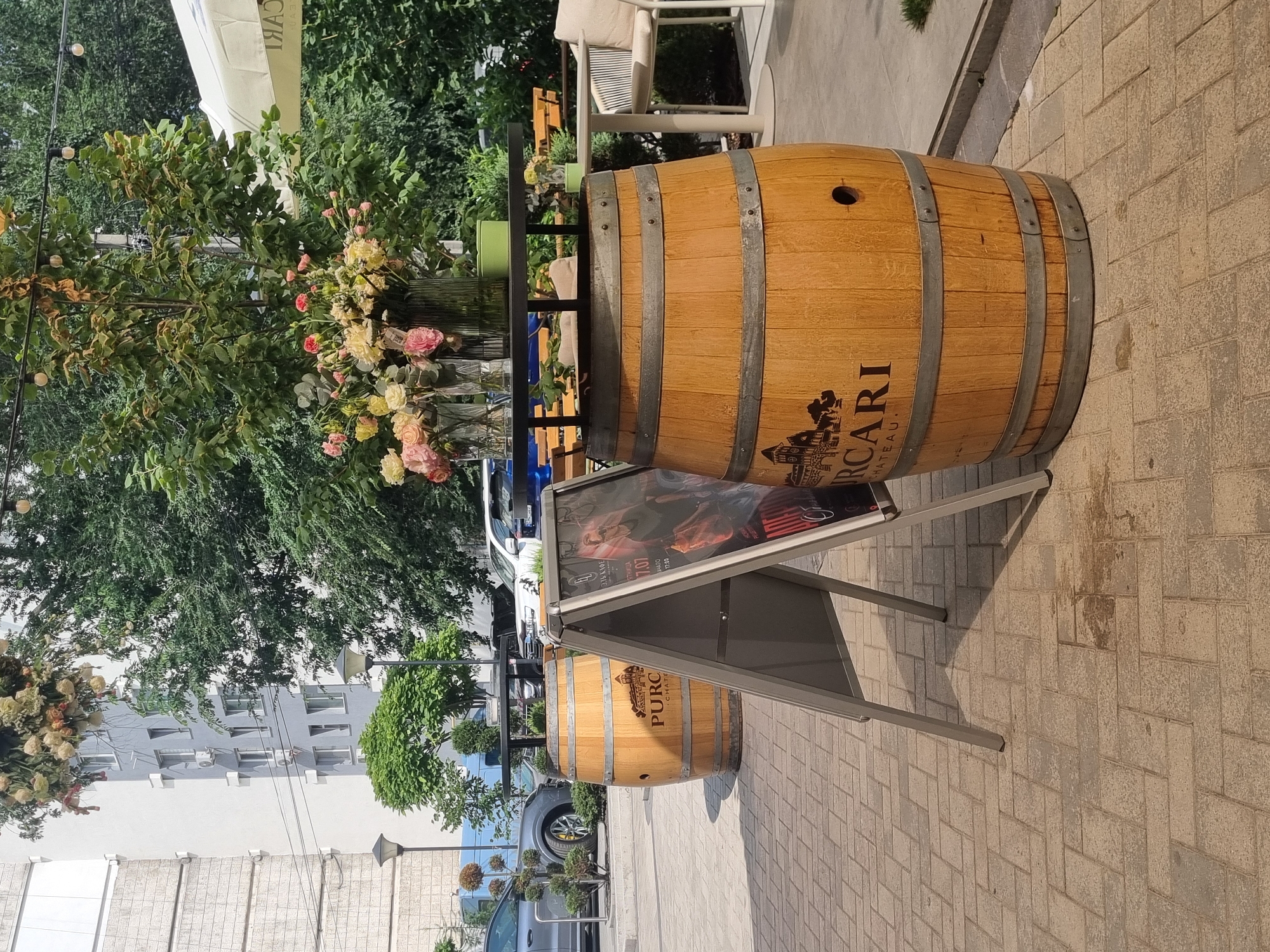 In an outdoor urban setting, a wooden wine barrel serves as a table, adorned with a large arrangement of colorful flowers in vases. Adjacent to the barrel, an A-frame sign displays a poster featuring a man in sunglasses and text in Cyrillic. The scene is situated on a paved walkway with a brick-like pattern. In the background, trees, a building with multiple windows, and parked cars suggest a commercial or residential area. A portion of a white umbrella is visible on the right side of the image.