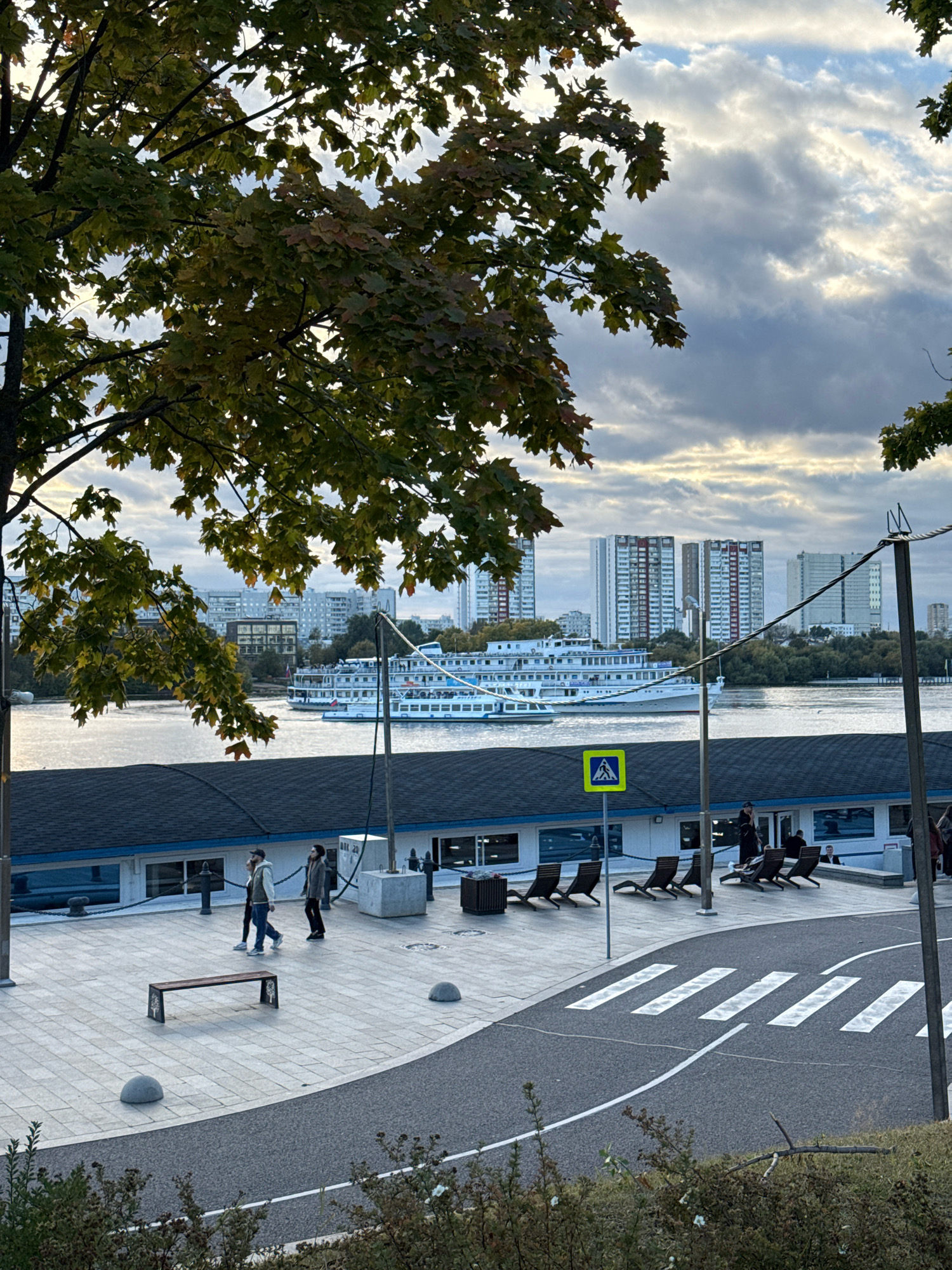 The image depicts a waterfront scene in Khimki, Russia, under a cloudy sky. In the foreground, a tree with autumn foliage partially obscures the view. A paved walkway with a curved asphalt road featuring a crosswalk runs across the bottom of the frame. Several people are visible on the walkway, including a group of three walking from left to right, and others seated or standing near a building with a dark roof and blue trim.
In the middle ground, a large white cruise ship is docked on a body of water, likely the Moscow Canal, given the location. The ship has multiple decks and windows, suggesting it is a passenger vessel. Across the water, a cityscape with numerous high-rise buildings is visible, characteristic of Khimki's urban landscape. A yellow pedestrian crossing sign is prominently displayed on the walkway.
The overall atmosphere is overcast, with diffused light. The scene captures a moment in an urban environment with a mix of natural elements, architecture, and human activity.