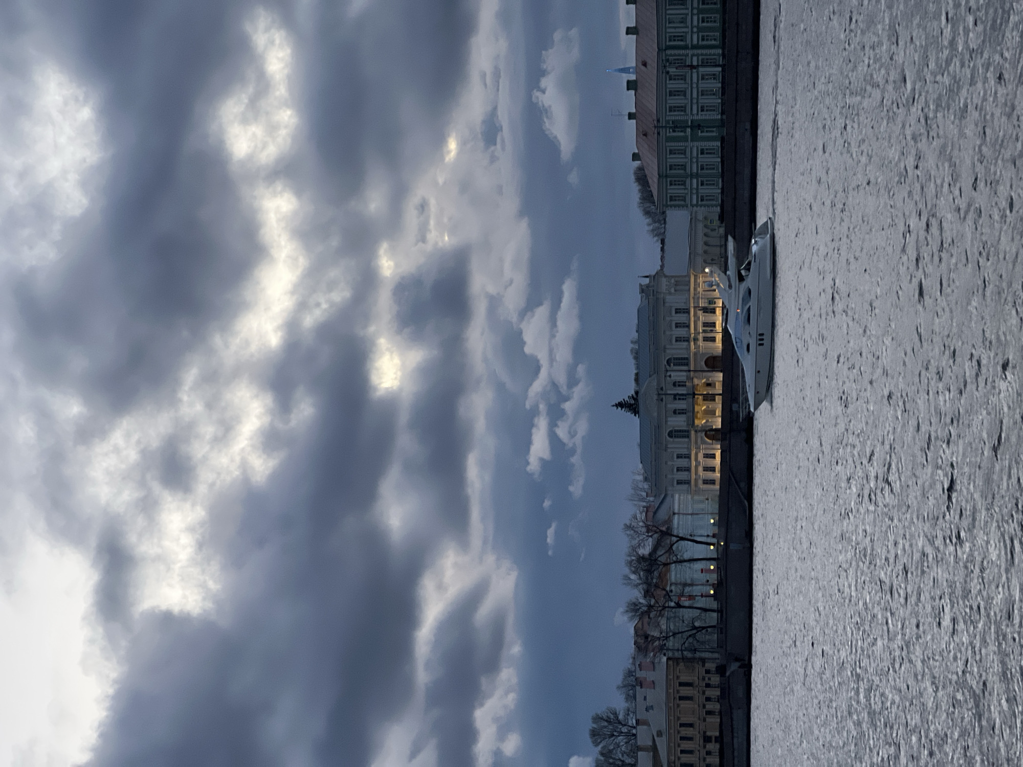 The image depicts a cityscape in Saint Petersburg, Russia, under a cloudy sky. A body of water, likely a river or canal, fills the foreground. A yacht is docked along the embankment. Buildings line the opposite bank. Streetlights are visible among bare trees. A prominent architectural feature is a building with a spire. The sky is overcast, with heavy clouds dominating the scene.