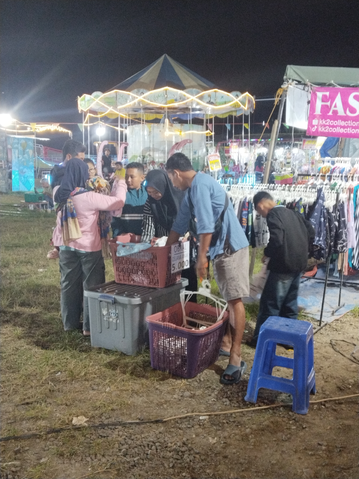 The image, taken at night, shows a group of people gathered around a clothing stall in Seloromo, Indonesia. The stall has a sign that says "OBRAL CELANA ANAK 5.000", with "OBRAL" appearing to be a clearance sale. Several baskets filled with clothes are in front of the stall, placed on a container. Several people are browsing the clothing. One person is wearing a blue shirt and khaki shorts. A woman in a pink shirt and hijab is carrying a child. Another man in a black jacket is examining items hanging on a rack. In the background, there is a carousel lit with yellow lights and other stalls. The ground is a mix of grass and bare earth. A blue plastic stool sits near the stall. A banner marked "FAS" is visible on another stall.