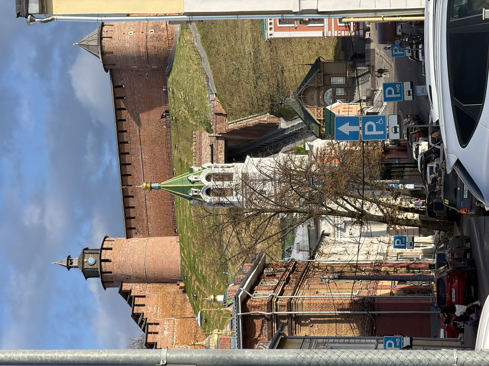 The image captures a view of the Nizhny Novgorod Kremlin from a street level. In the background, the imposing red brick walls of the Kremlin rise up a grassy embankment, with a prominent clock tower on the left and a conical-roofed tower on the right. A white bell tower with a green roof stands in the middle ground, and a steep staircase leads up towards the Kremlin walls.
In the foreground, the street is lined with buildings, some with red brick facades and ornate details. Several cars are parked and driving on the street, and a few pedestrians are visible. Traffic signs, including parking symbols and directional arrows, are placed along the roadside. The sky is partly cloudy with visible shadows cast by the sun. The overall scene depicts an urban environment with historical architecture.