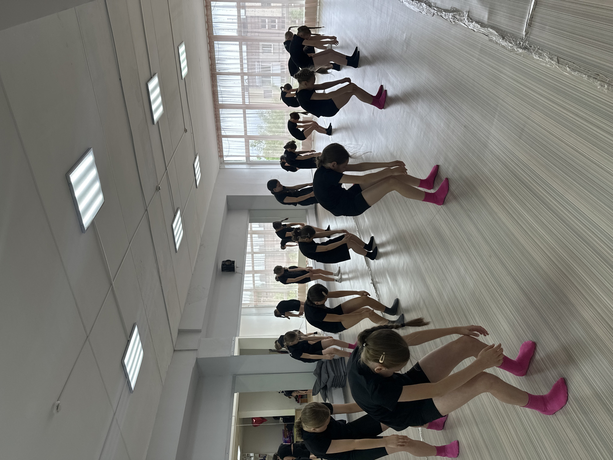 The image shows a dance studio in Votkinsk, Russia. Approximately 13 young girls are seated in rows on a light-colored, striped floor, possibly wood or linoleum. They wear black shorts and shirts; some have bright pink socks and others have black socks. They are bending forward, stretching their arms toward their legs. The room is well-lit with rectangular ceiling lights. There are windows with curtains in the background and a mirrored wall to the left, reflecting the room and some additional figures. The walls are white. The scene appears to be a dance or ballet class.