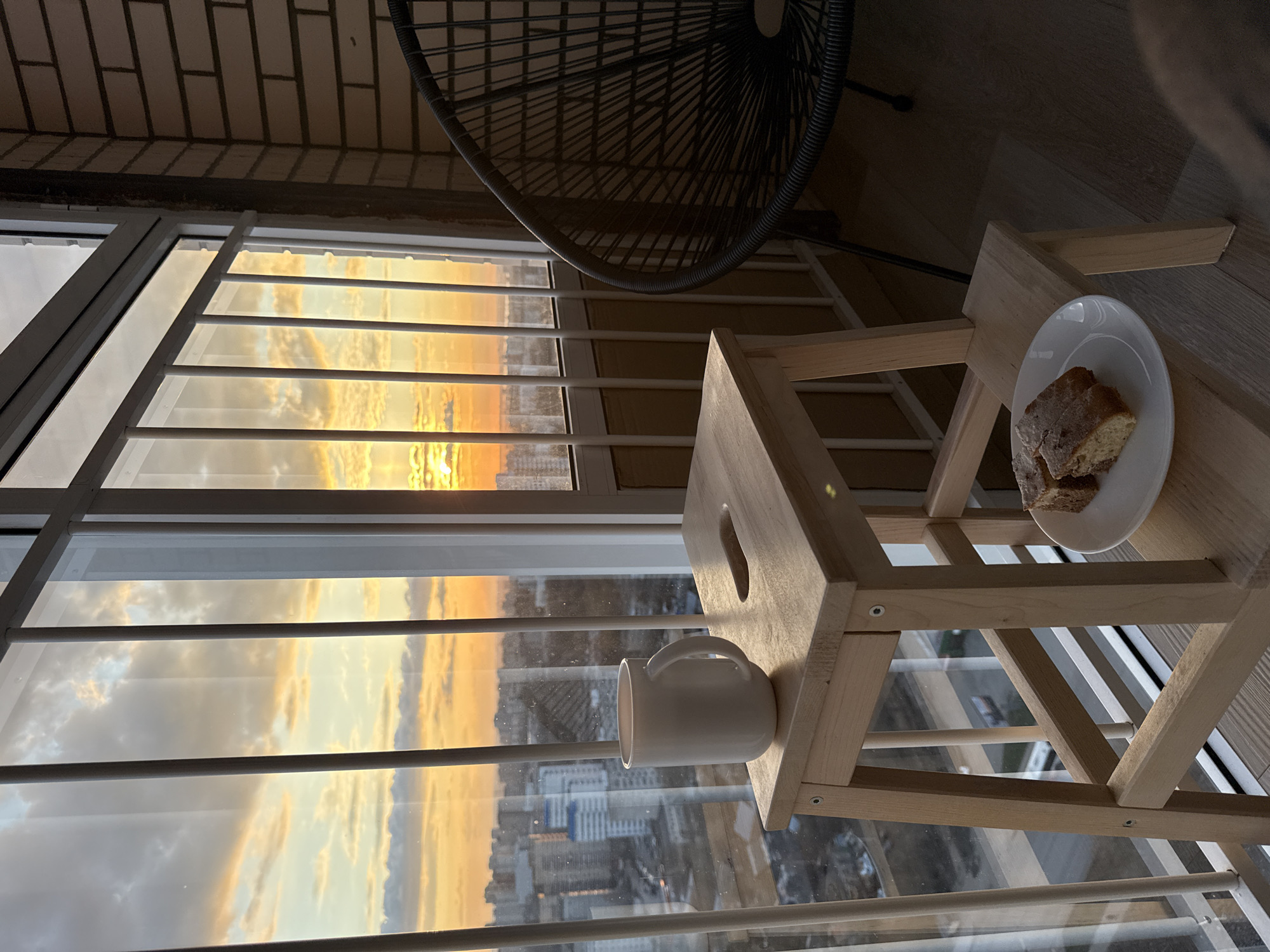 The image displays a tranquil scene from what appears to be an enclosed balcony or sunroom in Perm, Russia, during a vibrant sunset. The main focus is a large window offering a panoramic view of an urban landscape, bathed in the warm, intense hues of orange and yellow from the setting sun. Clouds in the sky are brilliantly illuminated, reflecting these colors.
In the foreground, a small, light-colored wooden stool or table is positioned by the window. On its lower shelf, a white ceramic mug rests, while on the upper surface, a white plate holds two pieces of cake or bread, suggesting a cozy moment for an evening snack. A section of a dark, woven wire-frame chair is visible in the upper right, hinting at a comfortable seating arrangement. The interior of the balcony features a brick-patterned ceiling and a wooden floor. The window structure, with its white frames and horizontal bars, reflects the golden light, adding to the warm ambiance.
Through the window, numerous city buildings can be seen stretching into the distance under the colorful sky. The scene evokes a sense of peace and relaxation. No people are visible, and there is no discernible text in the image. The time of day is clearly sunset, providing striking natural lighting.