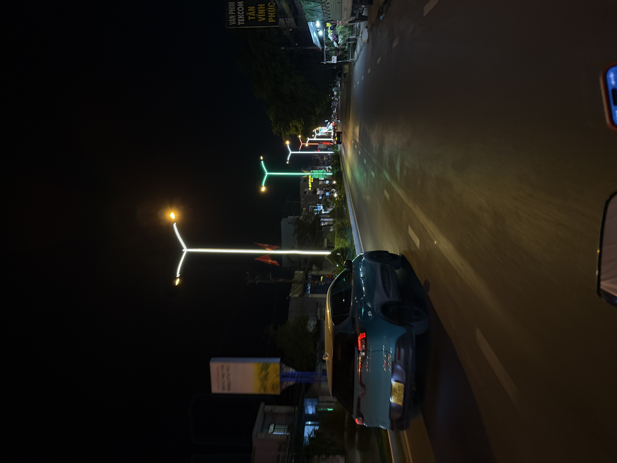 The image depicts a nighttime scene along a road in Diên Khánh, Vietnam. The view is likely from a moving vehicle, showing the roadway ahead on the right. A blue car is visible on the left side of the road. Above the car, there is a white rectangle sign. Overhead streetlights with a Y-shaped design illuminate the road and the surrounding area. Some flags, either national or municipal, can be seen in the background. A building is visible in the background of the image. There is also a street sign near the top of the image that has words on it.
