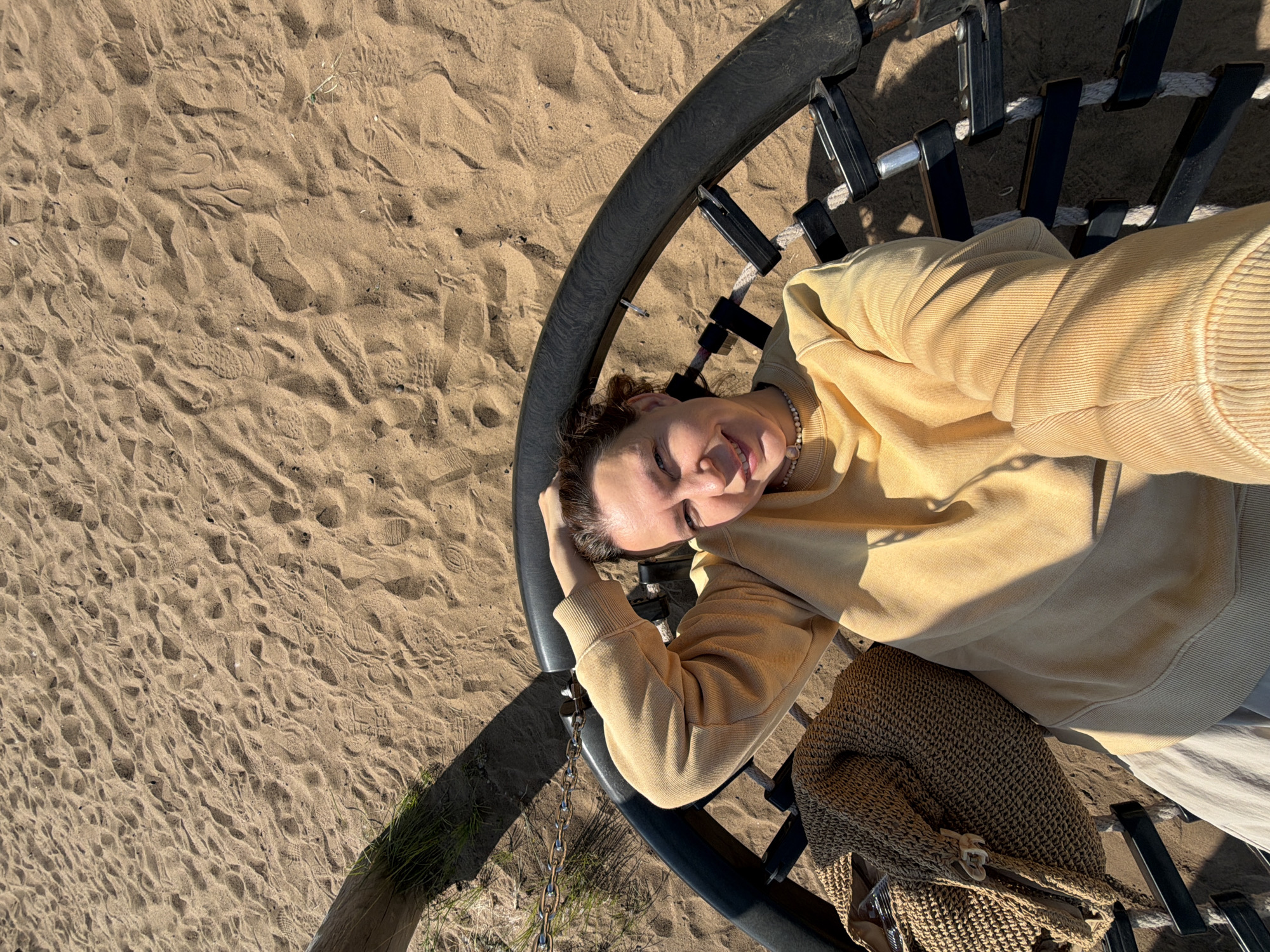 A person is lying on their back on a circular swing, with their head resting on their arm. They are wearing a yellow hoodie and a necklace. Next to them is a woven bag. The swing is suspended over a sandy area, with footprints visible in the sand. The background shows more sand and some dry grass. The lighting suggests it is daytime, with the sun casting shadows on the sand. The person appears to be smiling, conveying a relaxed and happy emotion. The location is identified as Chaikovsky, Russia, and the setting is likely a park or beach with a swing set. There is no visible text in the image.