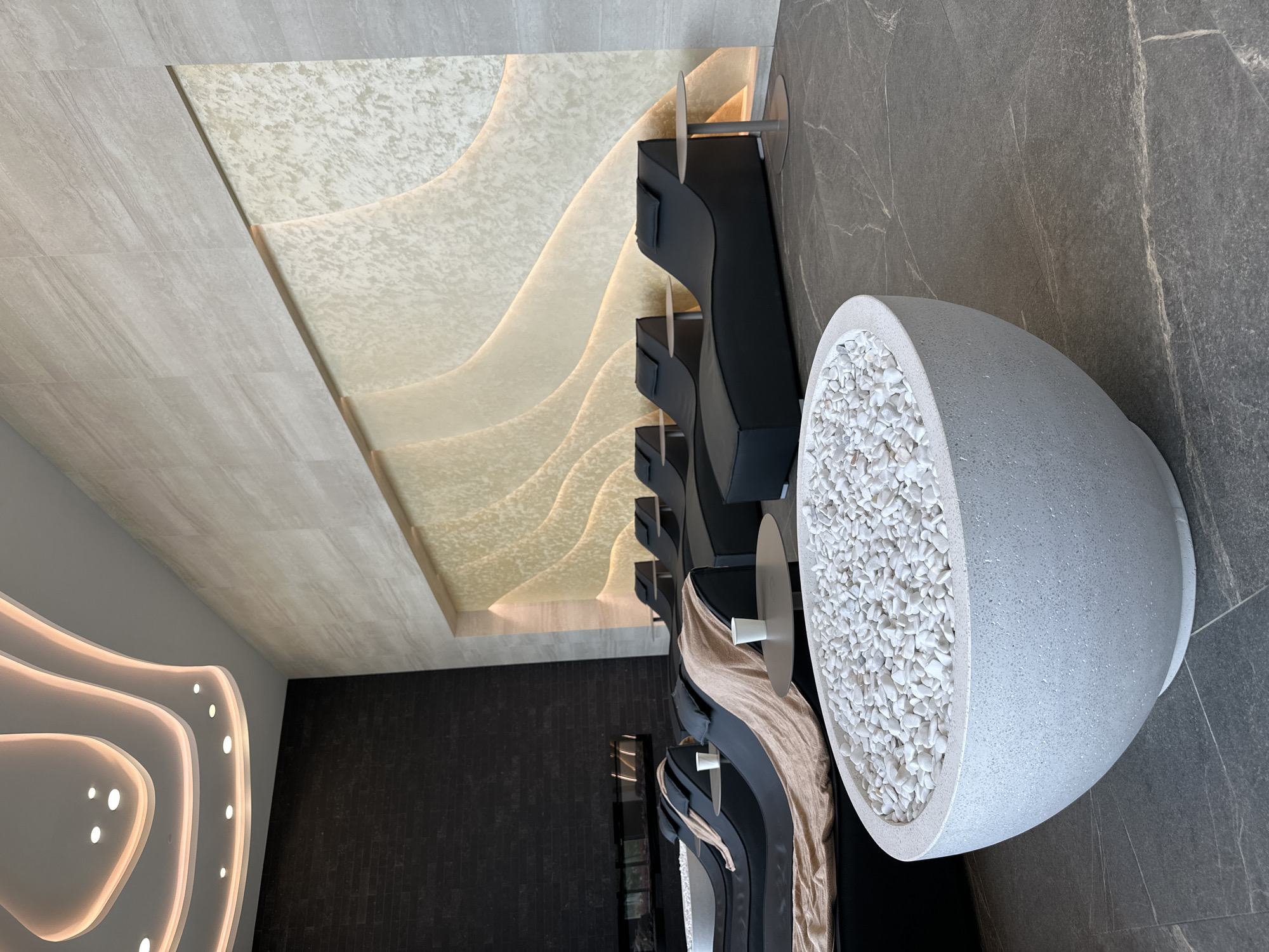 The image depicts an interior space, likely a lobby or waiting area. The location is Kazanlak, Bulgaria. The scene includes decorative architectural features, furniture, and flooring.
The ceiling has two distinct sections: one section features beige-colored textured panels with embedded lighting arranged in wave-like patterns, while the other part of the ceiling appears to be made of light grey tiles.
A series of black, modern-designed chairs or benches are visible, some with tan colored throws draped over them. Small round tables are placed adjacent to the seating.
A large, egg-shaped decorative vessel filled with white pebbles sits prominently in the foreground on a gray tiled floor. The walls behind are also covered in the same gray tiles, while a darker, textured black wall can be seen in the background.
No people are visible in the scene. The focus is on the architecture and interior design elements. The environment appears clean, modern, and aesthetically designed.