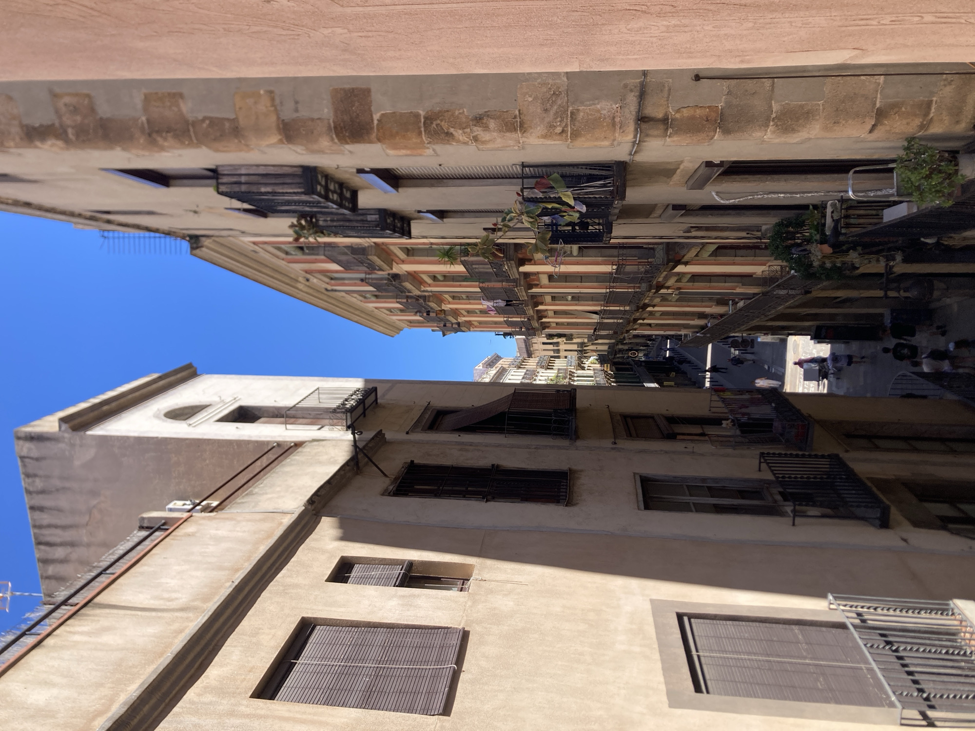 The image depicts a narrow street in Barcelona, Spain. Buildings with multiple stories line both sides of the street. The buildings are characterized by their old architecture, with visible windows, balconies, and decorative features. Some balconies are adorned with plants. The street is populated with pedestrians, suggesting a lively urban environment. The sky above is clear and blue.
