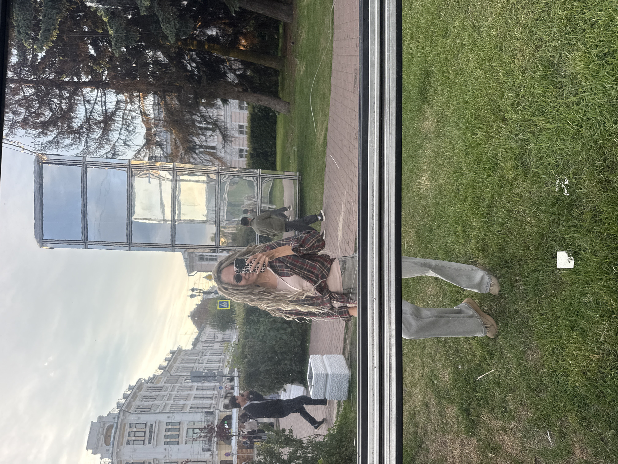 The media file shows a reflection selfie taken by a woman standing outdoors. Her upper body is reflected in a large, clear window or glass panel, occupying the upper left portion of the image. She has long, wavy blonde hair, wears sunglasses, a light-colored top, and an open plaid shirt (red, black, white). She is holding a smartphone with a white case that has unclear English text.
The reflection encompasses a city scene with a mix of modern glass buildings and older structures under a light, cloudy sky. Several other people are visible in the reflection, walking on a paved sidewalk. A prominent horizontal window frame divides the image. Below this frame, the woman's actual legs are visible, dressed in light-colored jeans and brown shoes, standing on green grass.
The outdoor environment includes a brick-paved sidewalk, lush green grass, and trees. The overall lighting suggests either late afternoon or early evening. A yellow diamond-shaped sign is also reflected in the window.