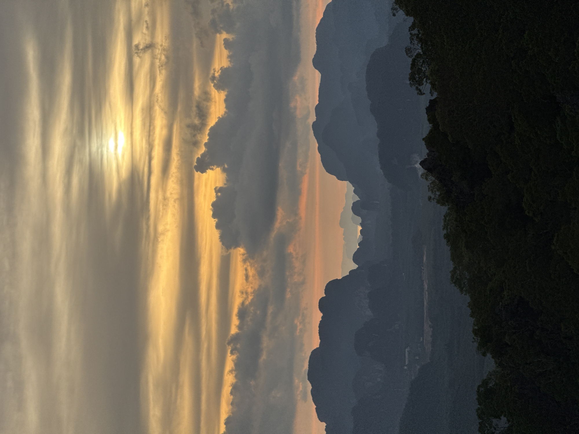 A wide-angle, low-angle shot captures a dramatic sunset over a mountainous landscape in Ban Khao Tang, Thailand. The sky is dominated by streaks of orange and yellow, with the sun a bright, hazy disc peeking through scattered clouds. Below the sky, a series of dark, silhouetted mountain ranges recede into the distance, their jagged peaks creating a stark contrast against the vibrant sky. The foreground is filled with the dark green silhouettes of trees and dense foliage, suggesting a forested area or a viewpoint overlooking a natural expanse. The overall impression is one of natural beauty and dramatic lighting.