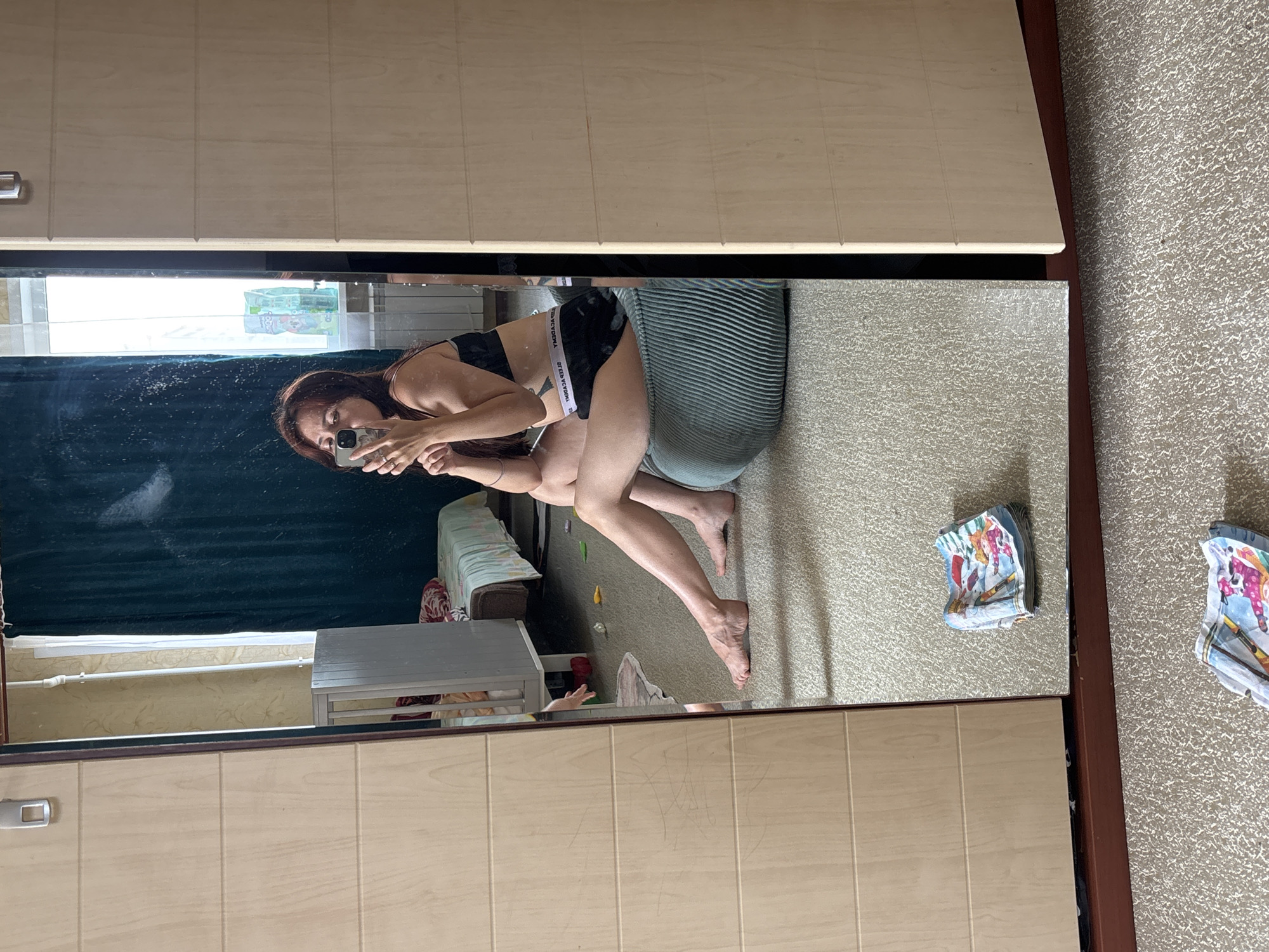 A woman is taking a selfie in a bedroom. She is sitting on a green ottoman, wearing black underwear and a black bra. Her legs are crossed and her right leg is extended. In the reflection of a mirrored wardrobe, a crib can be seen behind her, along with a window with curtains. A folded towel with a printed pattern is on the floor in front of the wardrobe. The room is carpeted. The mirrored wardrobe is light wood with horizontal panels.