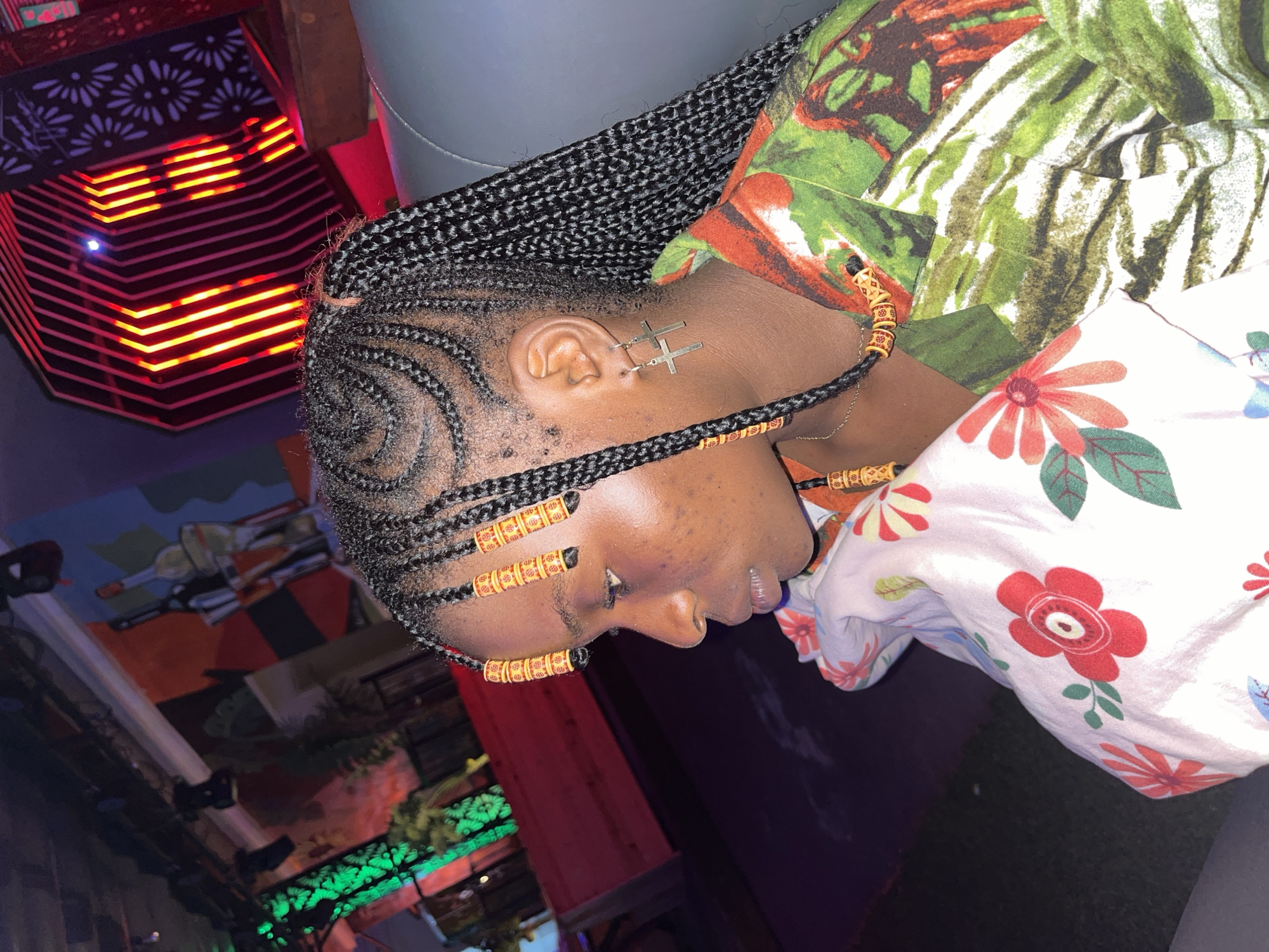 This image depicts a young Black woman in profile, seated in a dimly lit indoor establishment located in the city of Howayi, Nigeria. She faces left, with her gaze directed forward. Her hair is styled in intricate cornrows on the scalp, forming swirling patterns on the crown, and transitioning into individual box braids that extend down her back. Several braids framing her face are adorned with cylindrical yellow and orange beads featuring red patterns. A cross-shaped silver-colored earring hangs from her visible right ear, and a thin silver-colored chain necklace is visible around her neck, partially obscured by a beaded braid.
She wears a collared shirt with a vibrant, abstract, and possibly botanical pattern, primarily in shades of green, red, and orange. Her complexion shows minor blemishes on her cheek and forehead. She holds or rests against a white pillow or cushion patterned with large, stylized red and orange flowers and green leaves.
The background indicates a modern, leisure-oriented setting, likely a lounge or bar. To the left, blurred green lights suggest decorative foliage or illuminated panels. To the right, a prominent hexagonal structure with internal vertical red and orange lights serves as a striking decorative feature, next to a dark panel with a cutout pattern. Further in the background, dimly lit walls display abstract murals, including a depiction resembling bottles in an ice bucket. The overall ambiance is dark, illuminated by various colored accent lights.