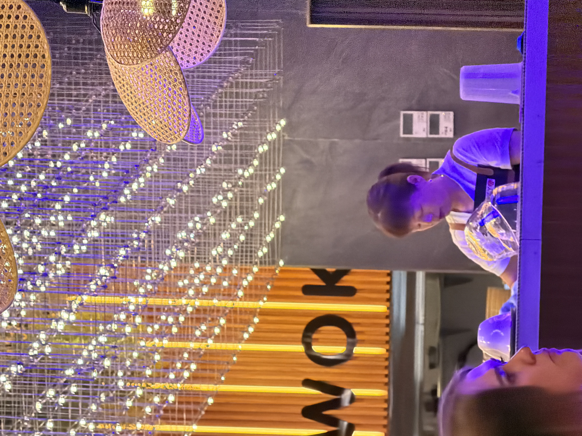 The image shows a vertical view of a dimly lit indoor space, possibly a restaurant or bar. The main subjects are two people, a woman and a man, and various decorative elements. The woman is in the foreground, her head and shoulders visible. She appears to be looking down, with a neutral expression. She is wearing a light-colored top. Further in the background, a person, presumably the man, is leaning over a counter. He is wearing a white t-shirt and has a lanyard around his neck. He is holding a glass containing a clear liquid.
The setting is characterized by vibrant lighting and decorative features. Hanging from the ceiling are woven light fixtures, possibly made of rattan, in circular and oval shapes. Behind the people, a grid-like structure is illuminated by numerous small, white lights, creating a sparkling effect. Below this, a wall is decorated with horizontal wooden slats, with large black letters spelling out "WORK". The lighting in the scene is a mix of warm yellow and cool purple tones, suggesting an evening or night setting. The overall atmosphere appears modern and atmospheric. There are no explicit weather cues, and the location is not identifiable beyond being indoors.