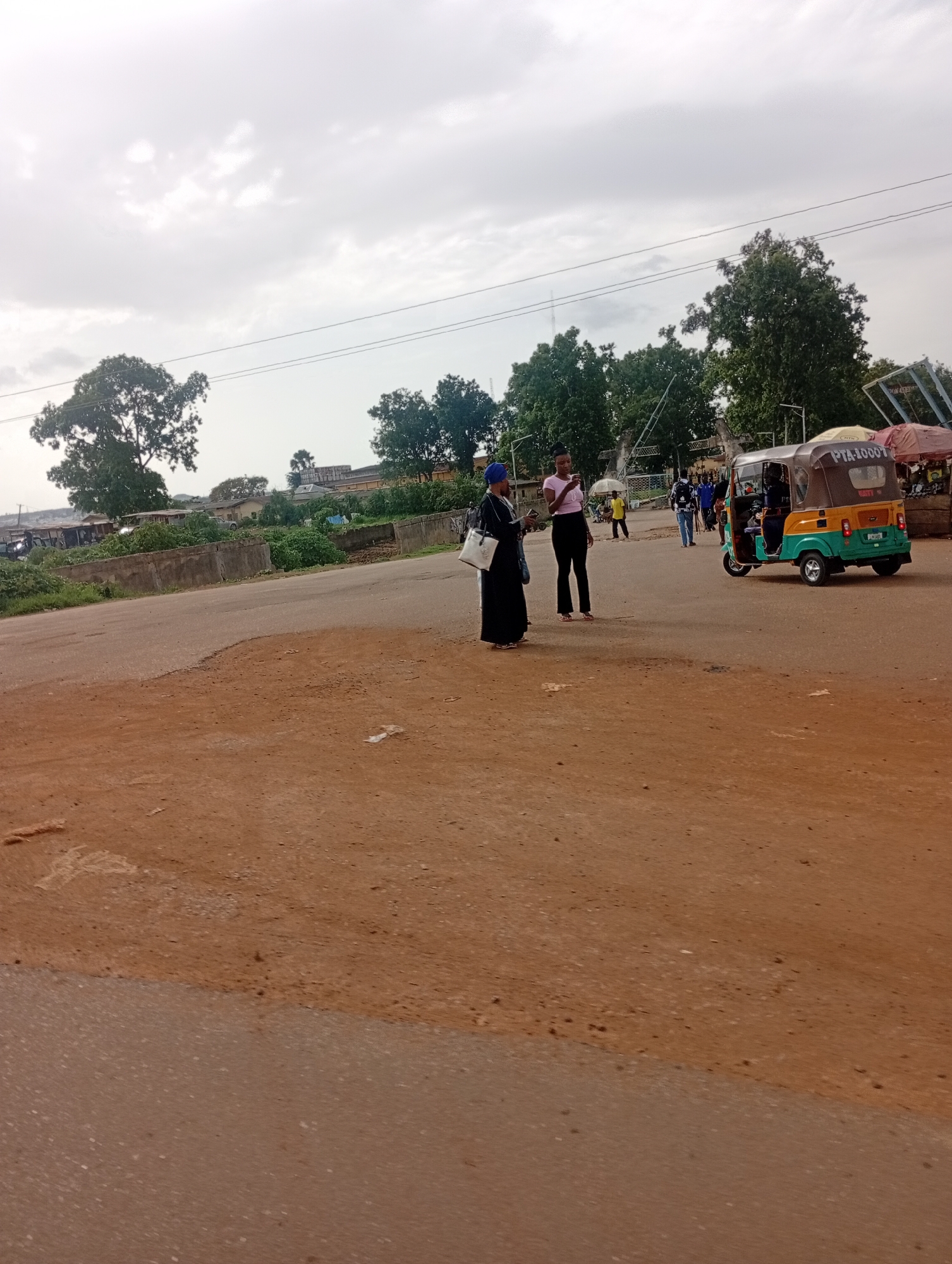 The image depicts a street scene in Rafin Par, Nigeria, on an overcast day. In the foreground, a dirt road transitions to asphalt. Two women are standing in the middle of the asphalt road. One woman, wearing a black abaya and blue headscarf, holds a white bag. The other woman, in a pink shirt and black pants, looks at her phone. Behind them, a yellow and green auto-rickshaw is parked, its door open. Several other people are visible in the background, along with trees and buildings. The overall impression is of a casual gathering in a somewhat dusty, semi-urban environment.