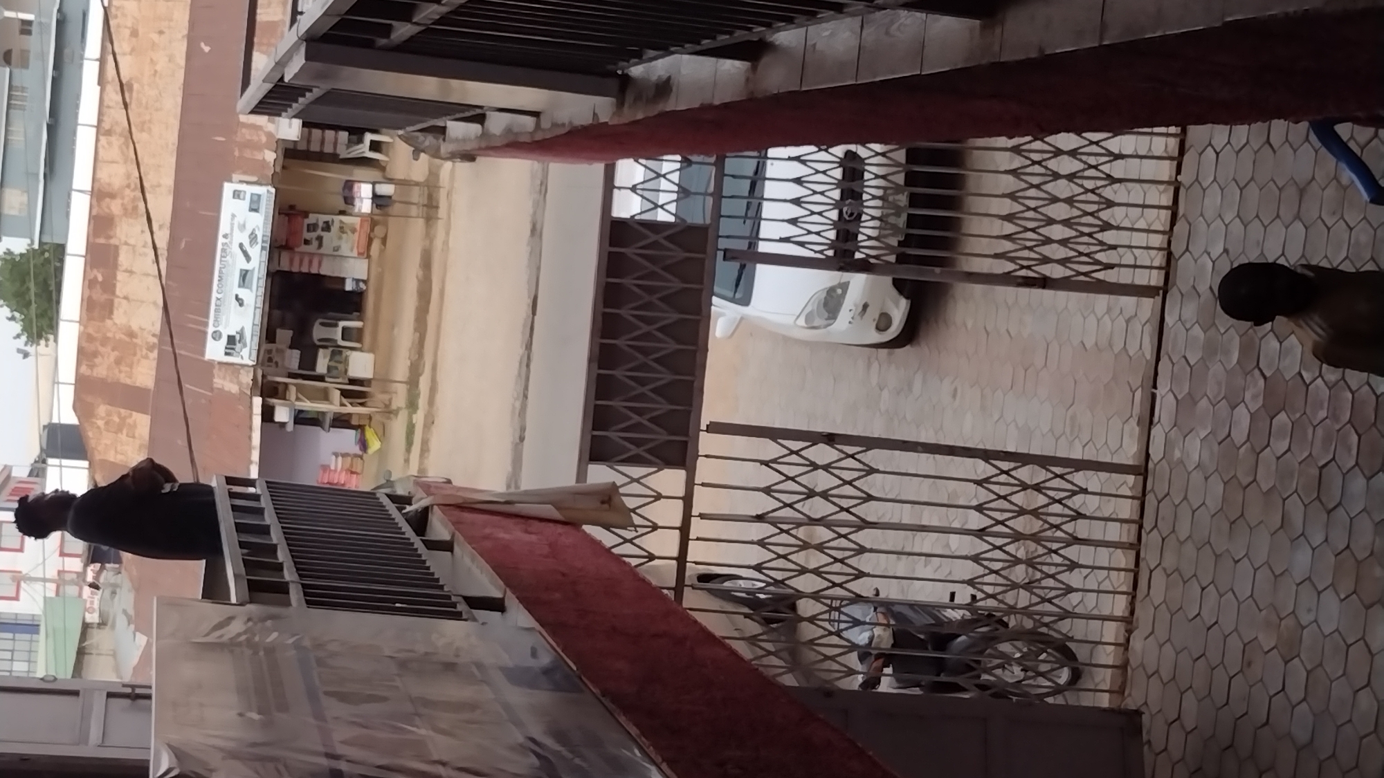 The image shows a street scene in Unknown, Unknown. From an elevated vantage point, a partial view of a building with what appears to be a computer store, "ORIBEX COMPUTERS &," is visible across the street. Parked on the street is a white car. A motorcycle is parked near a gated area. A person stands in the foreground of the elevated point. Another person is standing in the tiled area near the gated area. The scene is viewed through a metal lattice structure.