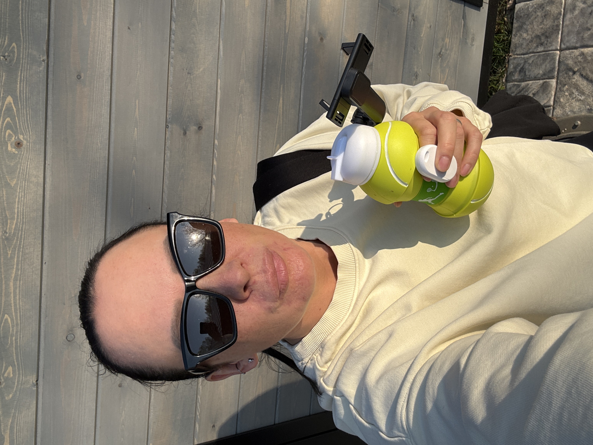 A person with dark hair and sunglasses is holding a yellow and white water bottle. The person is wearing a light-colored sweatshirt with a black collar underneath. A black phone mount is also visible. The background includes a gray wooden wall and paving stones. The location is unknown.