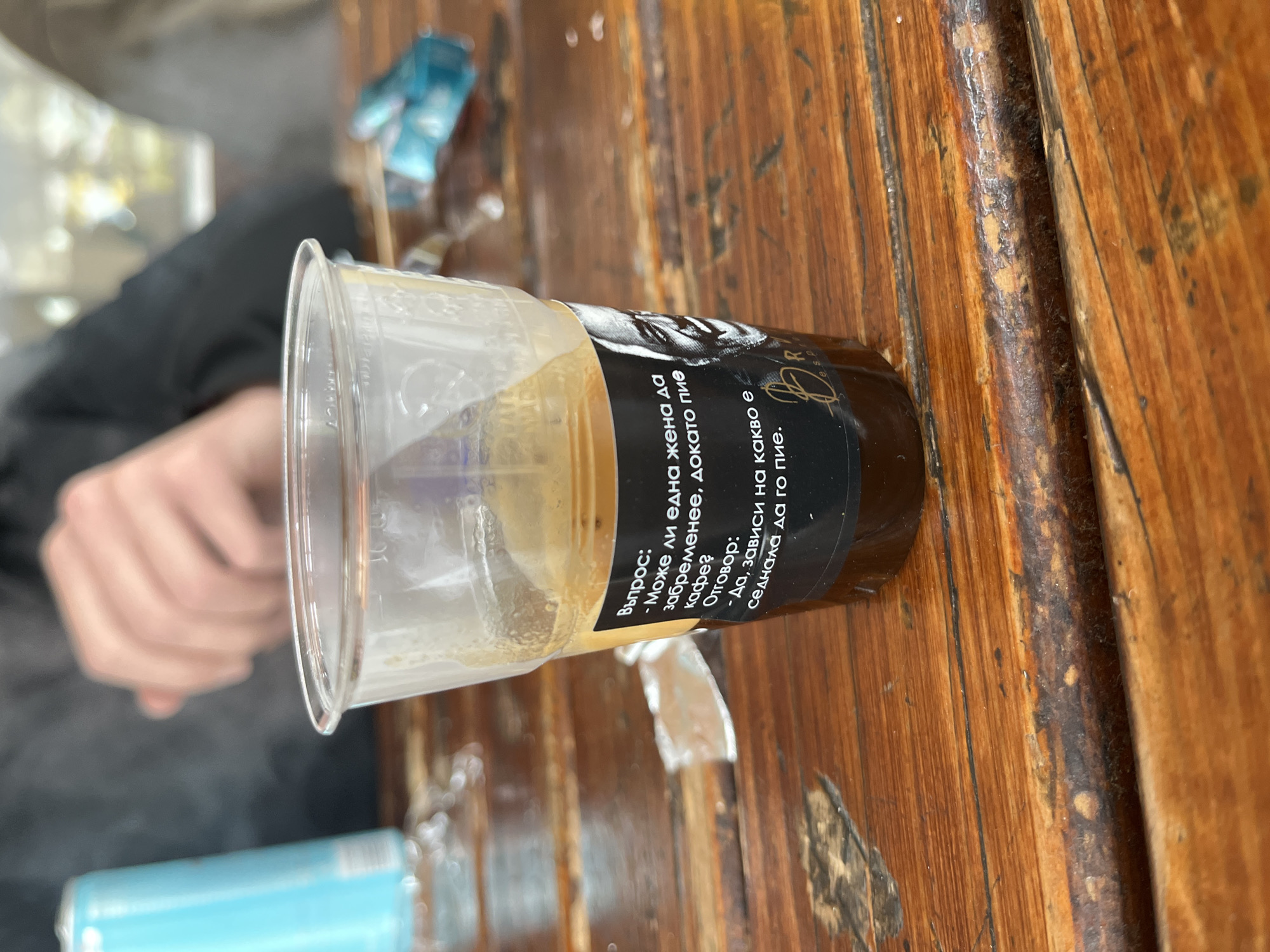 A plastic cup containing coffee sits on a wooden table. The cup has a black label with white text written in Bulgarian. The text translates to "Question: Can a woman get pregnant while drinking coffee? Answer: Yes, it depends on what she's sitting on to drink it." In the background, a person's hand can be seen, along with a blue can and some plastic packaging. The table is made of wooden planks with visible grain and wear. Based on the label being in Bulgarian, it is likely this image was taken in Bulgaria. However, more context is needed to confirm the specific location in Asenovgrad.