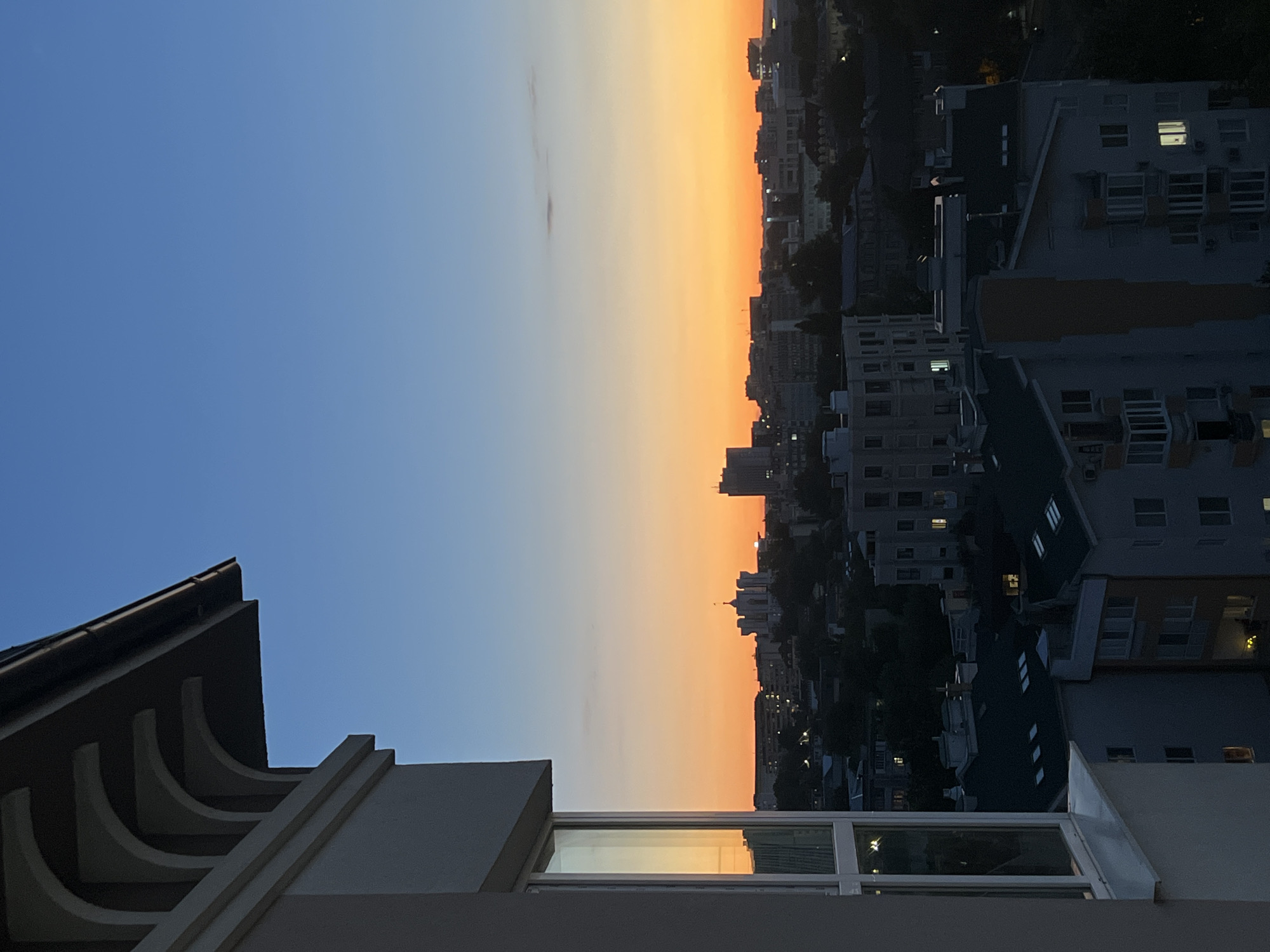 The image captures a twilight view of Chișinău, Moldova, from a high vantage point. In the foreground, the corner of a modern building with a white facade and a large window is visible, reflecting the warm hues of the sunset. The window frame is white, and the glass shows reflections of the sky and perhaps other buildings. Above this, a portion of the building's overhang is seen, featuring decorative, arched supports.
The middle ground offers a panoramic view of the city skyline as dusk settles. Various apartment buildings, characterized by their multi-story structures and dark roofs, are densely packed. Some buildings display illuminated windows, indicating ongoing activity within. A prominent white structure with a dome and spire stands out among the buildings in the distance, possibly a significant landmark. Other taller buildings, appearing more like office blocks or modern apartments, are also visible on the horizon. The cityscape is softened by the twilight and the silhouettes of trees interspersed among the structures.
The sky transitions from a deep blue at the top to a vibrant orange and yellow gradient near the horizon, a classic representation of sunset or sunrise. The overall atmosphere is serene and urban, showcasing the transition from day to night over the Moldovan capital. The image is a still photograph, with no discernible people or active events depicted.