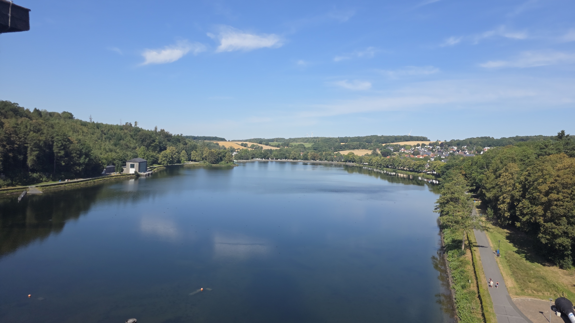 This is an aerial, wide-angle, daylight photograph of the Möhnesee in Möhnesee, Germany. The lake dominates the foreground and midground, reflecting a clear blue sky with sparse white clouds. The shoreline features a dense line of green trees. On the left bank, a small, modern, light-colored building with a dark roof is visible, adjacent to a forested hill. A paved path runs along the right bank, bordered by grass and trees. Several people are walking on this path. In the distance, rolling hills covered in greenery and agricultural fields are visible, along with a cluster of buildings forming a town. Wind turbines are present on the horizon. The overall scene depicts a serene, natural landscape with elements of human infrastructure and settlement.