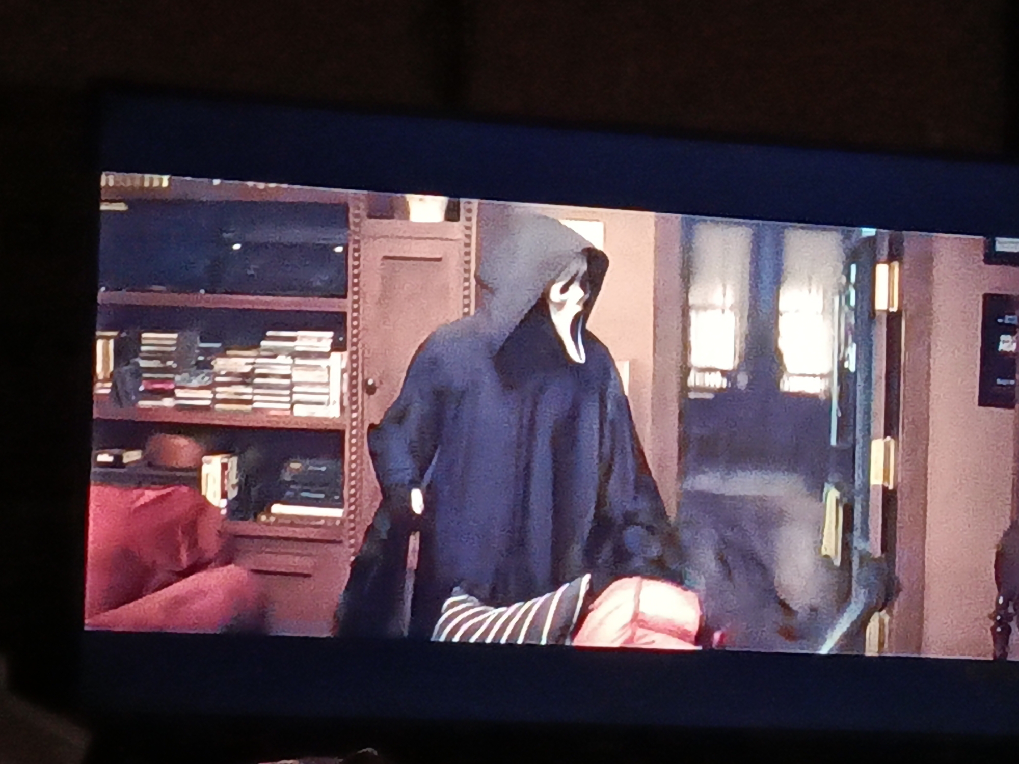 The image is a close-up shot of a television screen displaying a scene from the movie "Scream". The scene depicts the Ghostface killer standing in a room. Behind Ghostface is a dark wooden bookcase filled with books and objects. To the left is part of a red sofa. In the background are windows with light streaming through and part of a doorway. No information suggests it is Juan Griego, Venezuela.