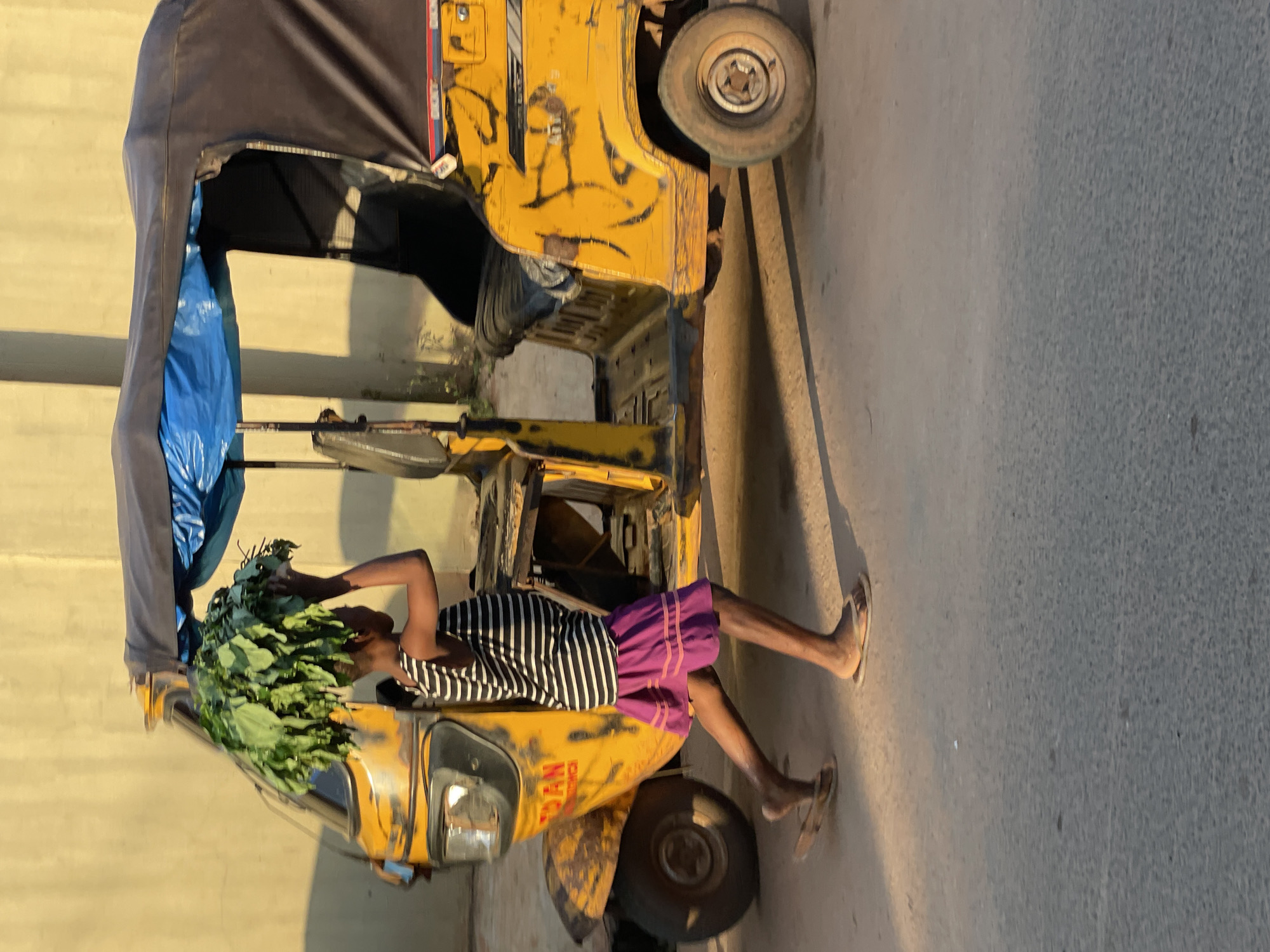 In Nkpor, Nigeria, a young person, likely a child, walks beside a yellow auto-rickshaw. The child, wearing a black and white striped sleeveless shirt and a purple skirt, carries a large bunch of green leafy vegetables balanced on their head. The auto-rickshaw, weathered and appearing somewhat worn, has a dark canopy over the passenger area. The scene is set on an asphalt road, with a pale yellow wall in the background, illuminated by what appears to be late afternoon sunlight casting shadows. The child is captured in motion, mid-stride, with their left leg extended forward. The vehicle's front is visible, as are its side panels, which bear some markings and what appear to be scratches or paint damage.