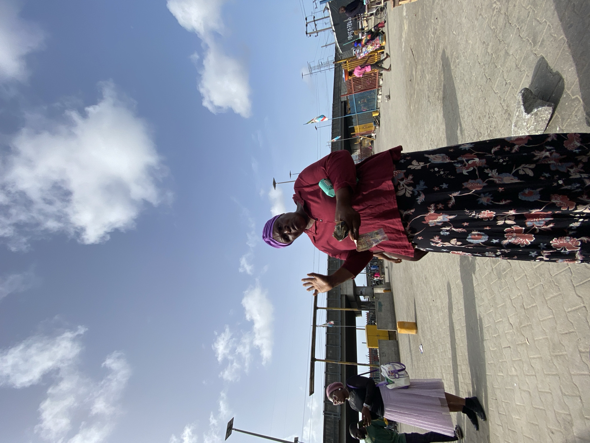 The image shows a woman in Ado, Nigeria. She is wearing a maroon top, a floral print skirt, and a purple head wrap. She is holding some bills in her left hand, and her right hand is raised. Behind her is a sky with clouds and an elevated roadway structure. Other people can be seen near the woman, including a woman with a light-colored skirt and a white bag, and a man in green clothing. The ground is paved. In the background are utility poles and commercial structures.