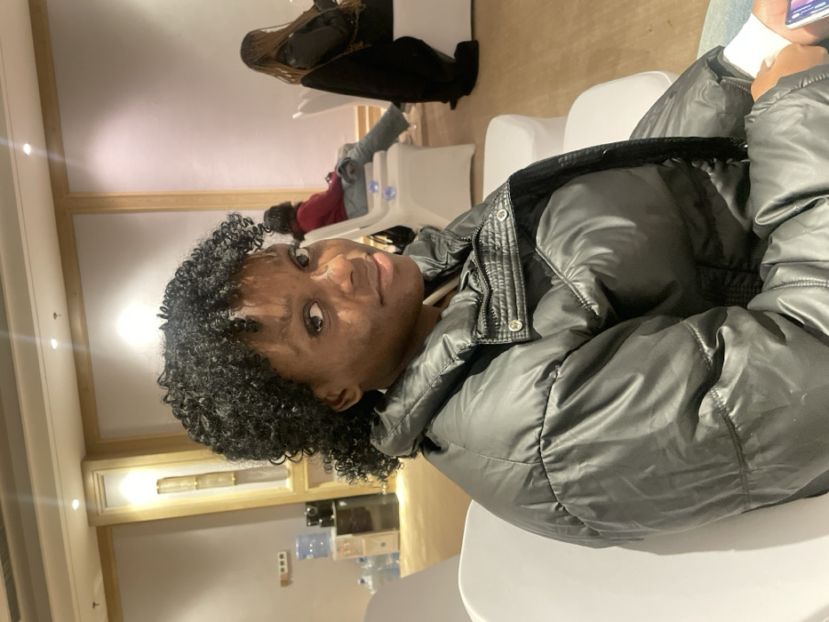 A person with curly black hair, wearing a black puffer jacket, sits in a white chair and looks directly at the camera. They appear to be indoors, possibly in a waiting area with white walls and decorative lighting. Other people are seated in the background, and a cabinet or display with various objects sits against the wall. The scene appears somewhat informal and brightly lit.