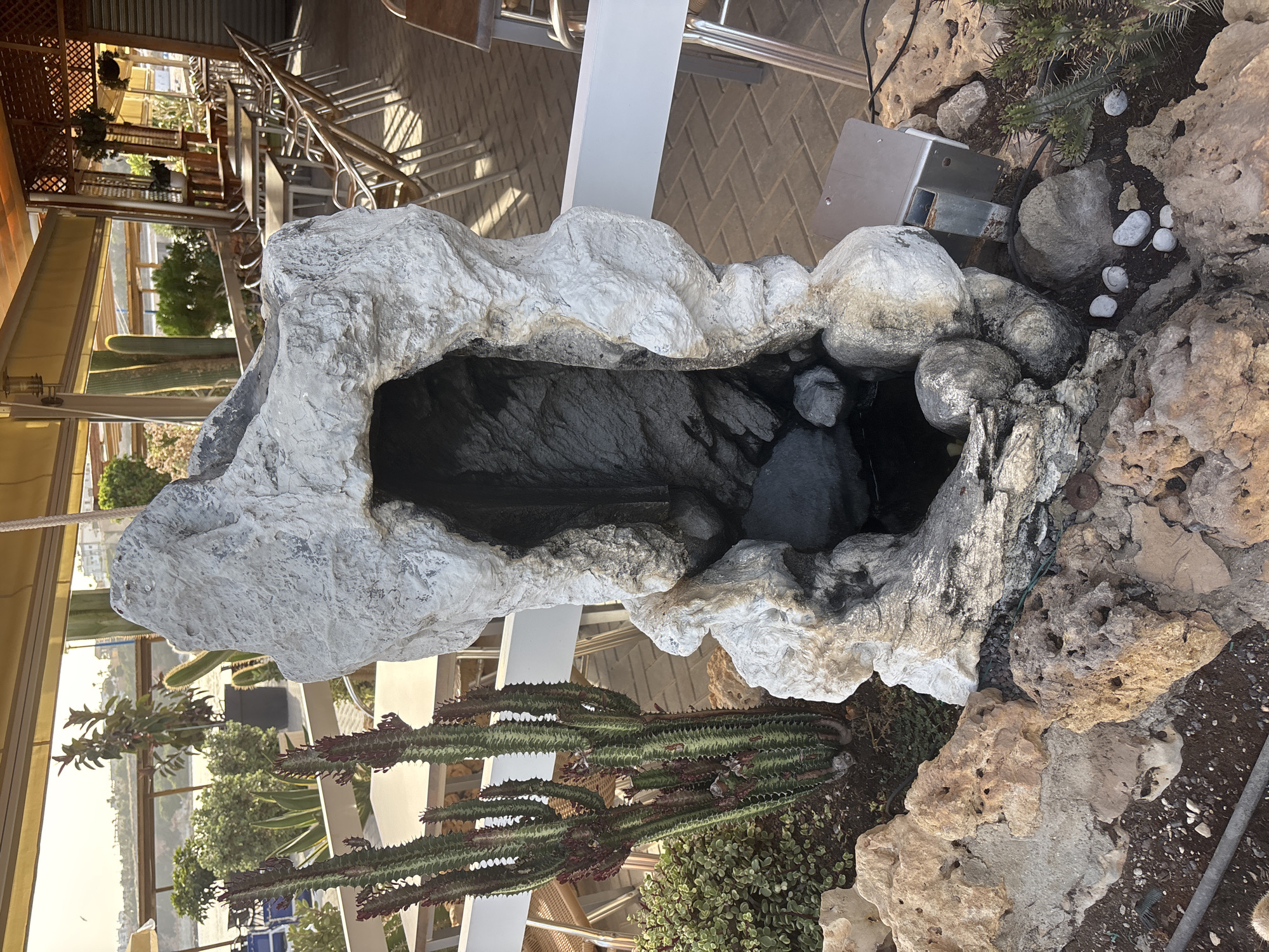 The image depicts an outdoor seating area with tables and chairs, likely a cafe or restaurant, in Orihuela, Spain. In the foreground, a large, artificial rock formation resembling a cave or grotto is prominently displayed. A tall, spiky cactus stands to the left of the rock feature. In the background, several tables and chairs are set up, some covered by awnings. Beyond the seating area, a body of water and some distant buildings are visible, suggesting a coastal or waterside location. The overall atmosphere appears to be relaxed and Mediterranean, with natural light illuminating the scene.