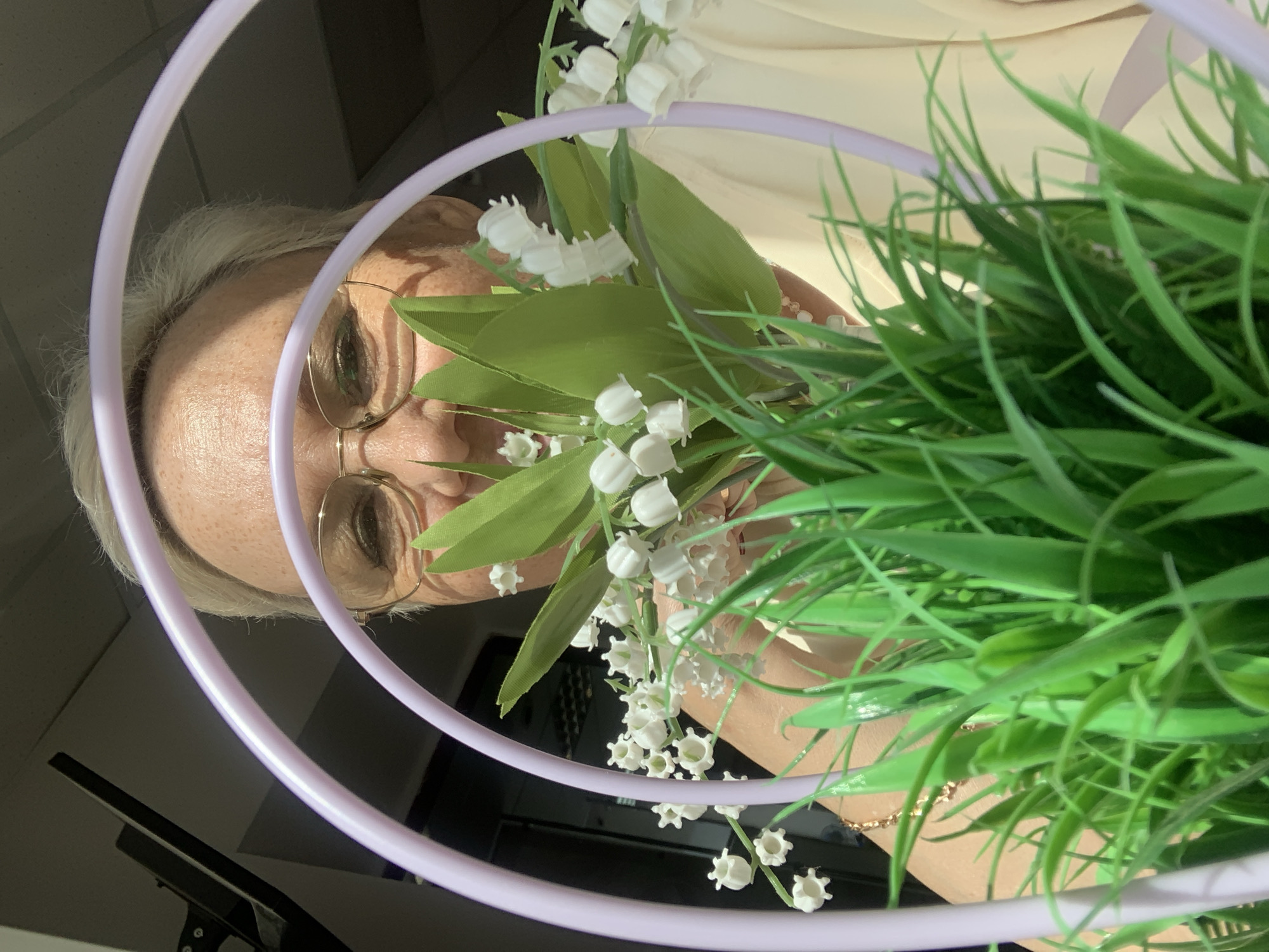 A close-up, portrait-oriented image features an older woman partially framed by decorative elements and artificial plants. The woman, with short, light-colored hair and wearing gold-rimmed glasses, is visible from her forehead to her upper arm on the left side of the frame. She has fair skin and appears to be looking forward, with a neutral expression.
She is holding or arranging a bouquet of artificial white lily of the valley flowers with green leaves, which occupy the central part of the image. To the right, a dense bunch of vibrant green artificial grass is prominent. Two concentric, glossy light purple hoops diagonally traverse the scene, creating a circular frame around the woman's head and the floral arrangements. A delicate gold-colored chain or bracelet is visible on her right wrist.
The background behind the woman and plants is indistinct and light-colored. In the lower left of the image, a dark, reflective surface shows a clear reflection of what appears to be a black monitor or screen with a stand, suggesting an indoor environment such as a home or office. The lighting is soft and even, indicating an indoor setting. No specific time of day or weather conditions are discernible, and there is no visible text. The image likely captures a moment of arrangement or posing with artificial floral decorations in Chaikovsky, Russia.
