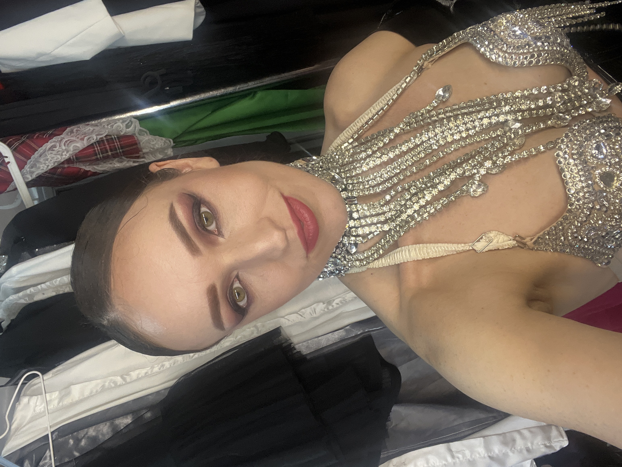 An image shows a woman with dark hair pulled back, wearing dramatic makeup including red lipstick and smoky eyeshadow. She is adorned with a sparkly, multi-layered rhinestone necklace and a matching bra-like garment. The background appears to be a closet or dressing room, with various garments hanging on a rack. The lighting is somewhat dim, and the photo is taken from a close-up perspective, focusing on the woman's face and upper body.