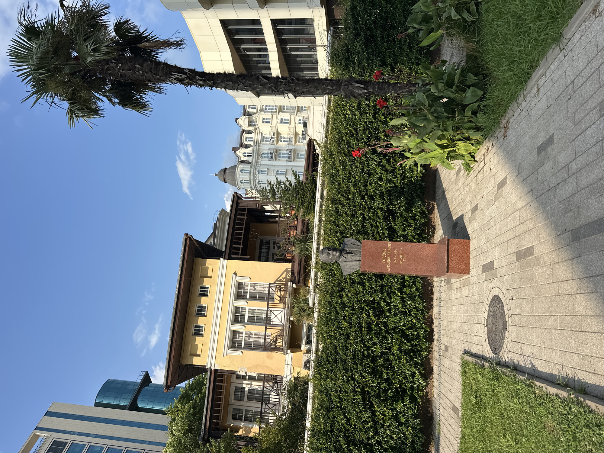 This image depicts a bust of Arkady Ignatievich Gorzon, the first doctor of Sochi, located in a park-like setting with modern and historic buildings in the background. The bust is made of bronze and rests on a red granite pedestal inscribed with his name and dates of life (1871-1940). The pedestal is situated in front of a neatly trimmed green hedge.
To the left of the bust, a paved walkway with a circular manhole cover is visible. To the right, a tall palm tree stands next to a modern building with a curved facade. In the mid-ground, a two-story yellow house with balconies and a dark roof stands out, while in the background, several multi-story buildings with classical architecture and a tall, modern glass-and-steel skyscraper are visible. Red flowers and lush green plants add color to the foreground. The sky is clear blue with a few wispy clouds. The scene suggests a sunny day in a well-maintained urban area.