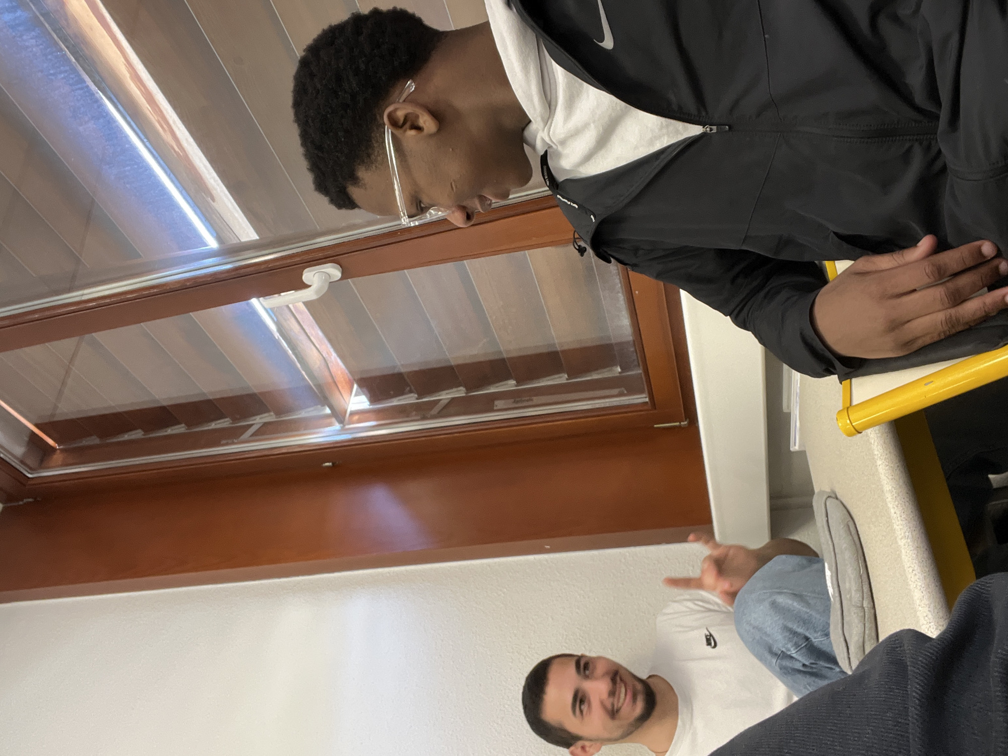 The image shows two young men inside a building in Schiltigheim, France. One is wearing a black and white Nike jacket and clear-framed glasses. The other is wearing a white t-shirt and giving a hand gesture. The scene includes a window with blinds, a desk with a yellow edge, and other clothing items. The building's interior is characterized by white walls and brown window frames.