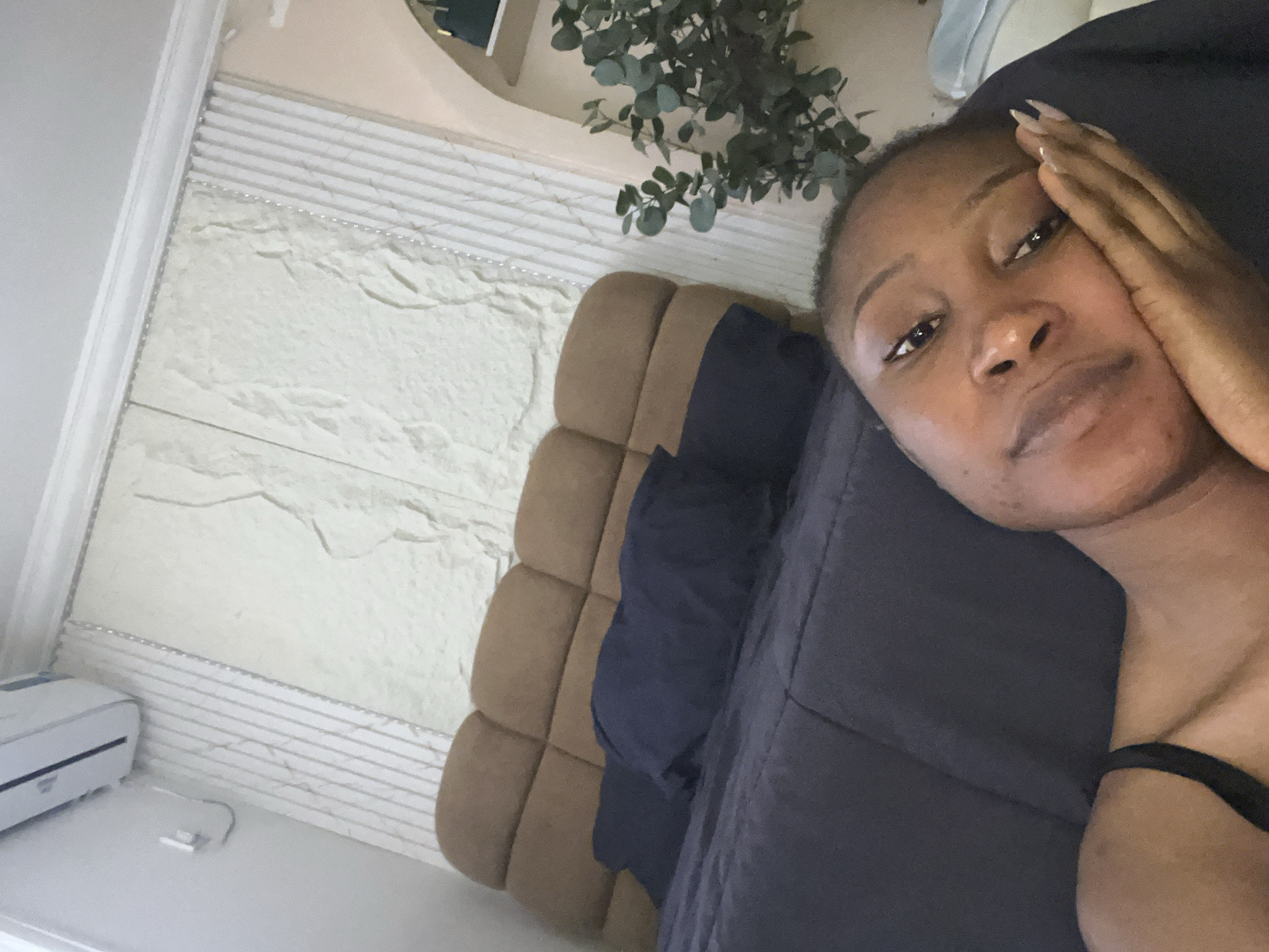 A woman is lying on a bed, with her face close to the camera, supported by her hand. She is wearing a black top. The background shows a bed with a brown headboard, navy blue pillows, and a dark blue duvet. To the right of the bed, there is a wall with decorative panels and a shelf with a green plant. An air conditioning unit is mounted on the wall above the headboard. The scene appears to be an indoor bedroom setting, likely in Abuja, Nigeria.