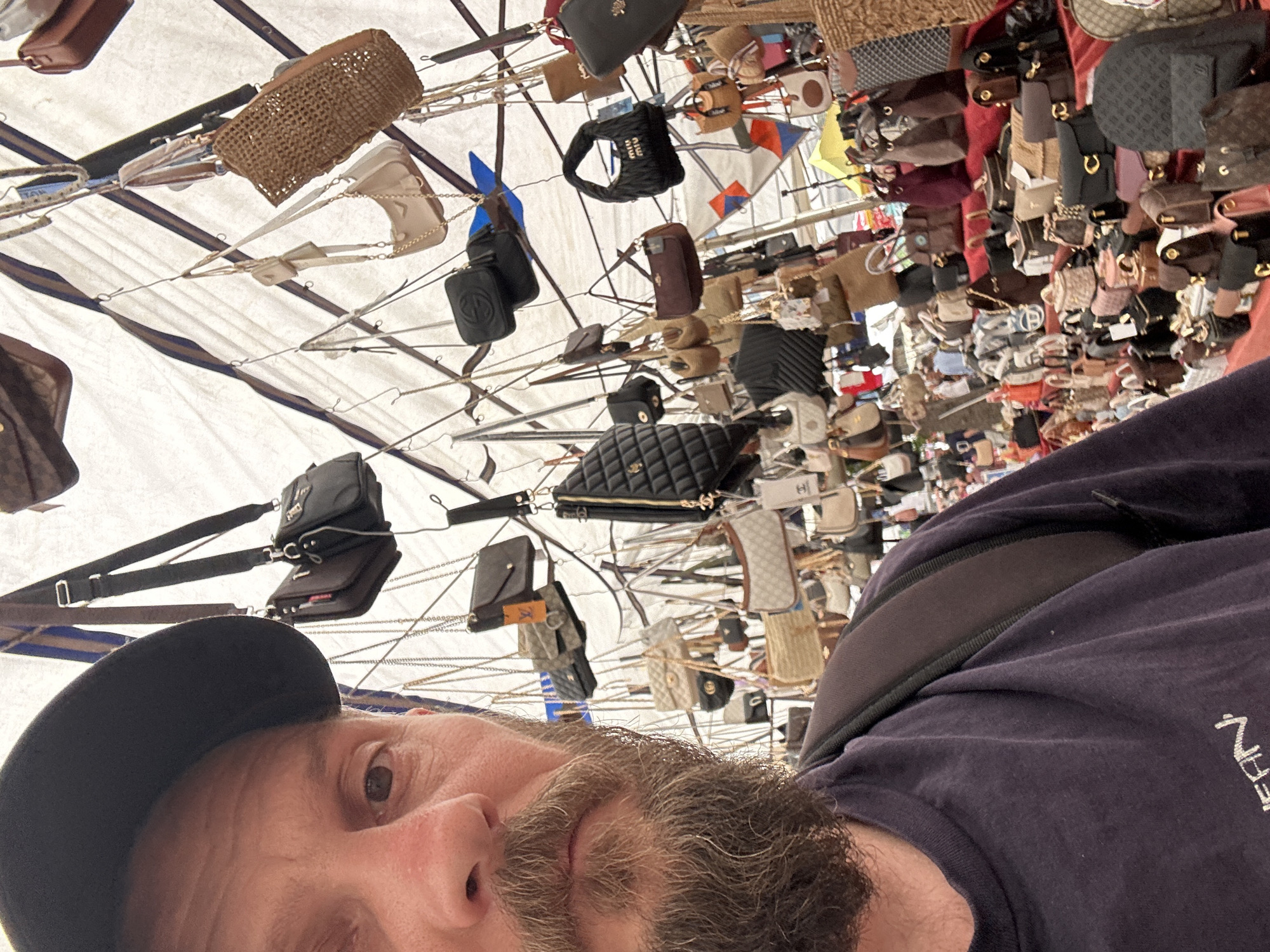 The image is a selfie of a Caucasian man with a beard, wearing a black baseball cap and a dark blue t-shirt with white lettering on the right side. He is standing under a canopy covered with numerous hanging handbags and accessories. The variety of items suggests the location is likely a market or bazaar. Based on the prompt mentioning Fethiye, Turkey, it is plausible that this scene is taking place within the Fethiye market. The items overhead, ranging from woven bags to luxury-branded items, are displayed for sale. The overall environment appears busy and crowded.