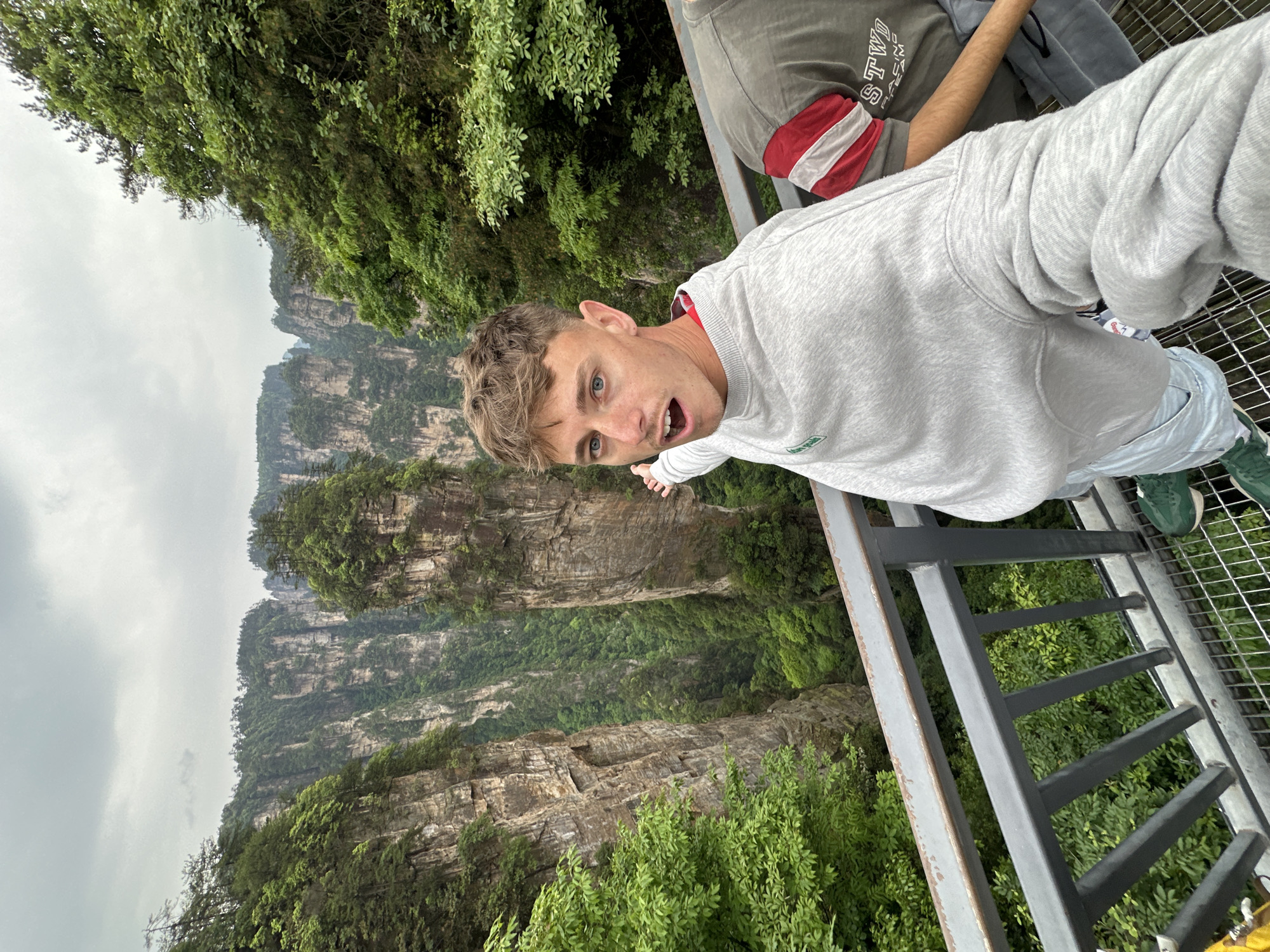 The image captures a selfie taken by a young man with short brown hair and blue eyes, positioned on a metal grate platform overlooking a mountainous landscape. The man wears a gray sweatshirt, light blue pants, and green shoes. He appears to be leaning over a metal railing, with an open mouth expression and a slightly twisted body position. A second person partially appears in the background, wearing a gray shirt with a red and white stripe on the sleeve.
The environment reveals a dramatic mountainous backdrop with lush green vegetation clinging to the rock faces. The sky is overcast and gray. The scene is likely set in the Tianmenshan National Forest Park of Luoguta, China.