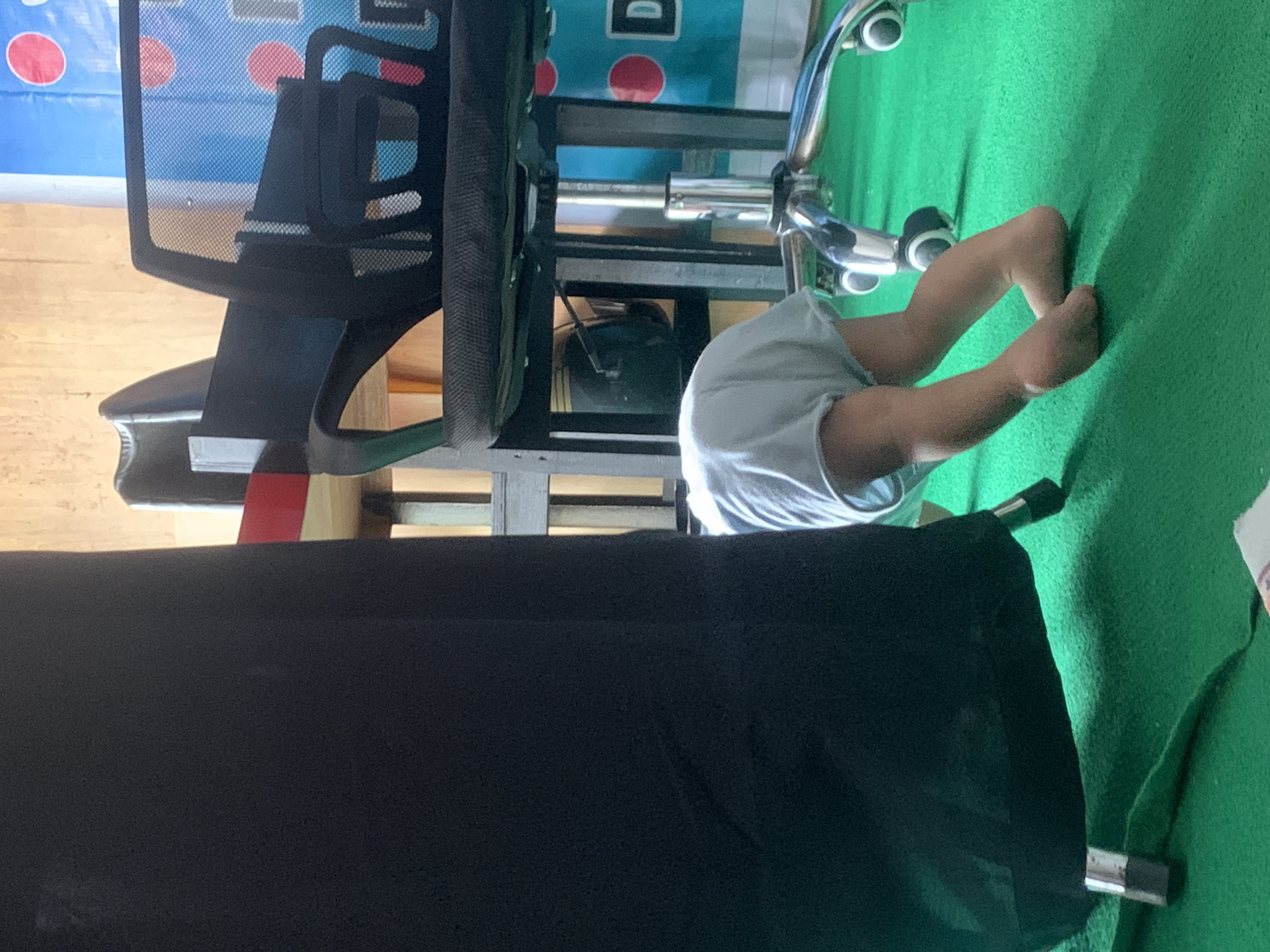 A child, wearing a light blue diaper, is on all fours on a green carpet. The child's head is bent downwards, obscured by a large black object that appears to be a speaker or cabinet. Behind the child, part of an office chair with a mesh back and chrome accents is visible. The background shows a wooden wall and a blue banner with red circles and the letter "D". The scene appears to be indoors, possibly in a room in Sungai Meriam, Indonesia, given the context provided.