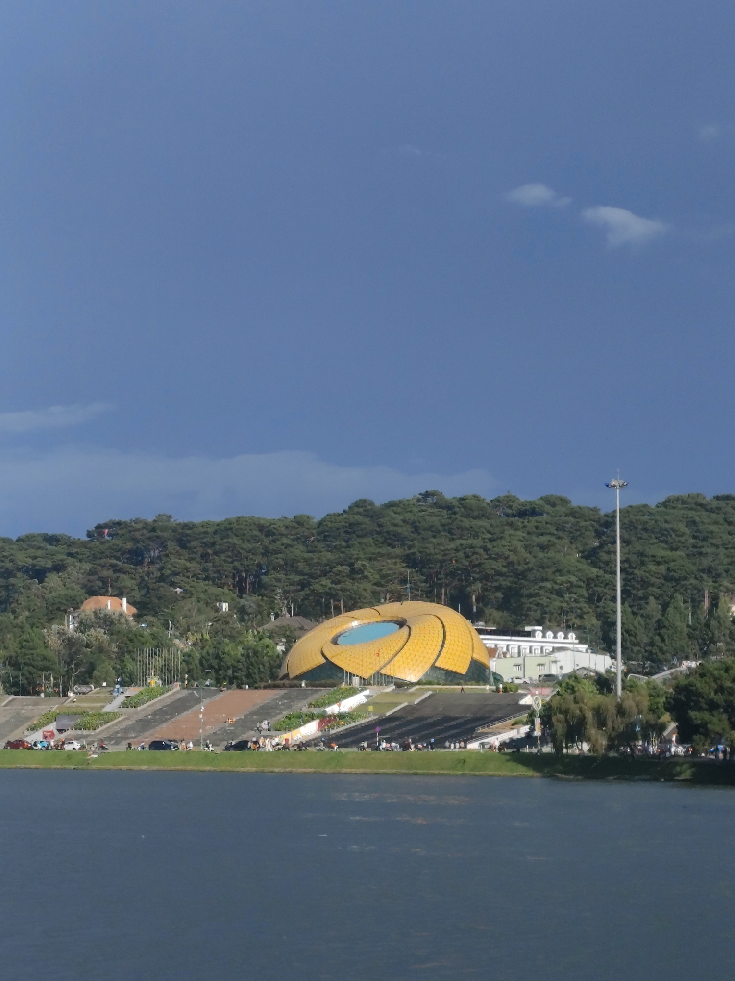 The image shows a wide, calm body of water in the foreground, likely Xuan Huong Lake in Da Lat, Vietnam. Beyond the water, a landscaped park area with tiered seating and pathways stretches towards a distinctive, bright yellow, lotus-shaped building. The building features a blue circular element at its center. Behind the building, a dense, green pine forest covers a hillside. Several vehicles, including cars and motorcycles, are parked or driving along the pathways in front of the tiered seating area, where numerous people are visible. A tall lamppost stands to the right of the yellow building, and a few buildings with white facades are nestled amongst the trees on the hillside. The sky above is a deep, cloudy blue, with a few lighter clouds present.