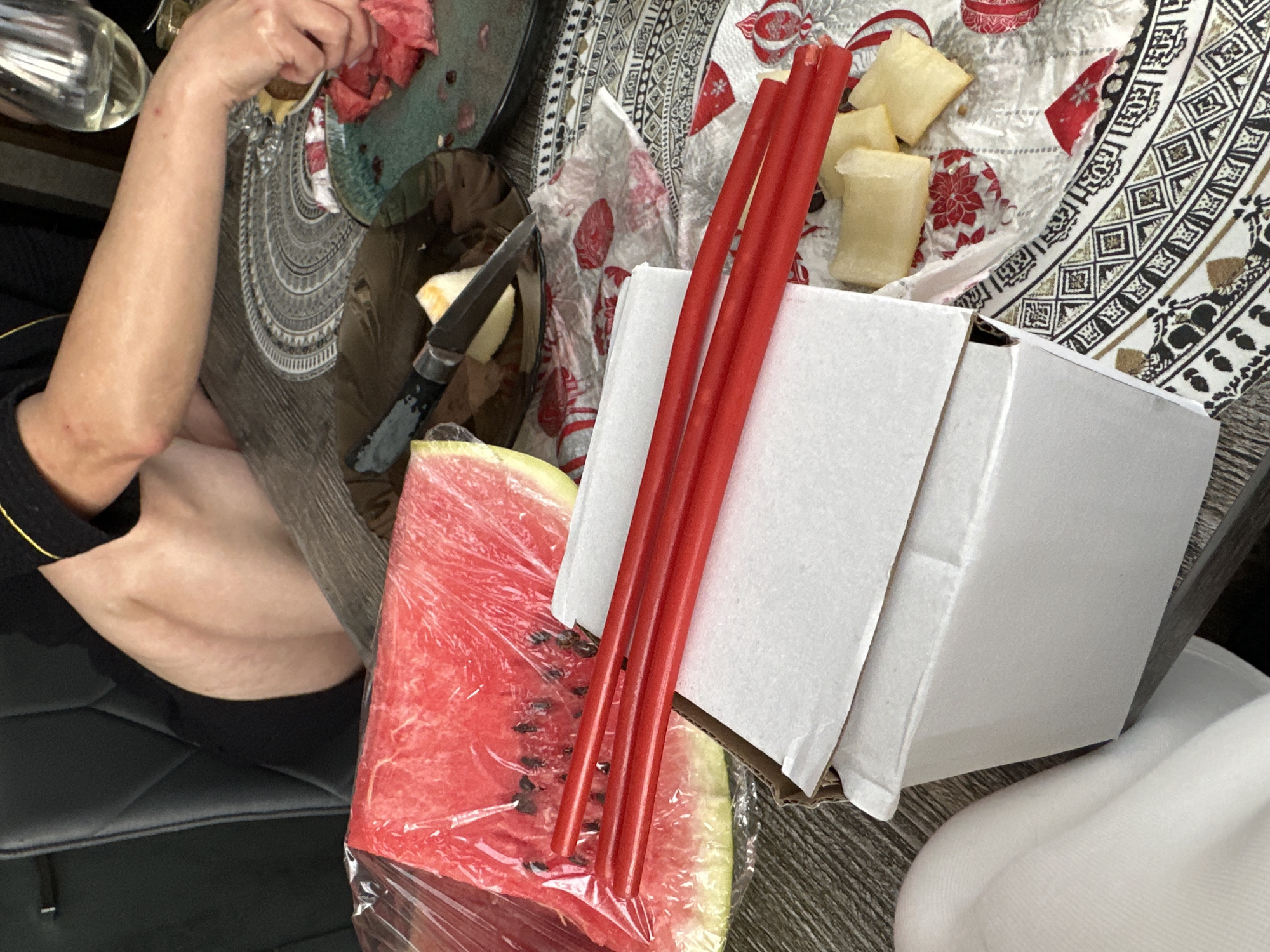 The image depicts a close-up, overhead view of a table setting with a person's arms and legs visible, suggesting they are seated. On the table, a large slice of watermelon is wrapped in plastic. Next to it, a stack of white boxes is placed, with three red chopsticks resting on top. In the background, a partially eaten plate of watermelon, a knife, and a wine glass are visible. The table is covered with a patterned tablecloth. The scene appears to be indoors, likely a dining area. The specific location in Baklashi, Russia is not evident from the visual information provided in the image.