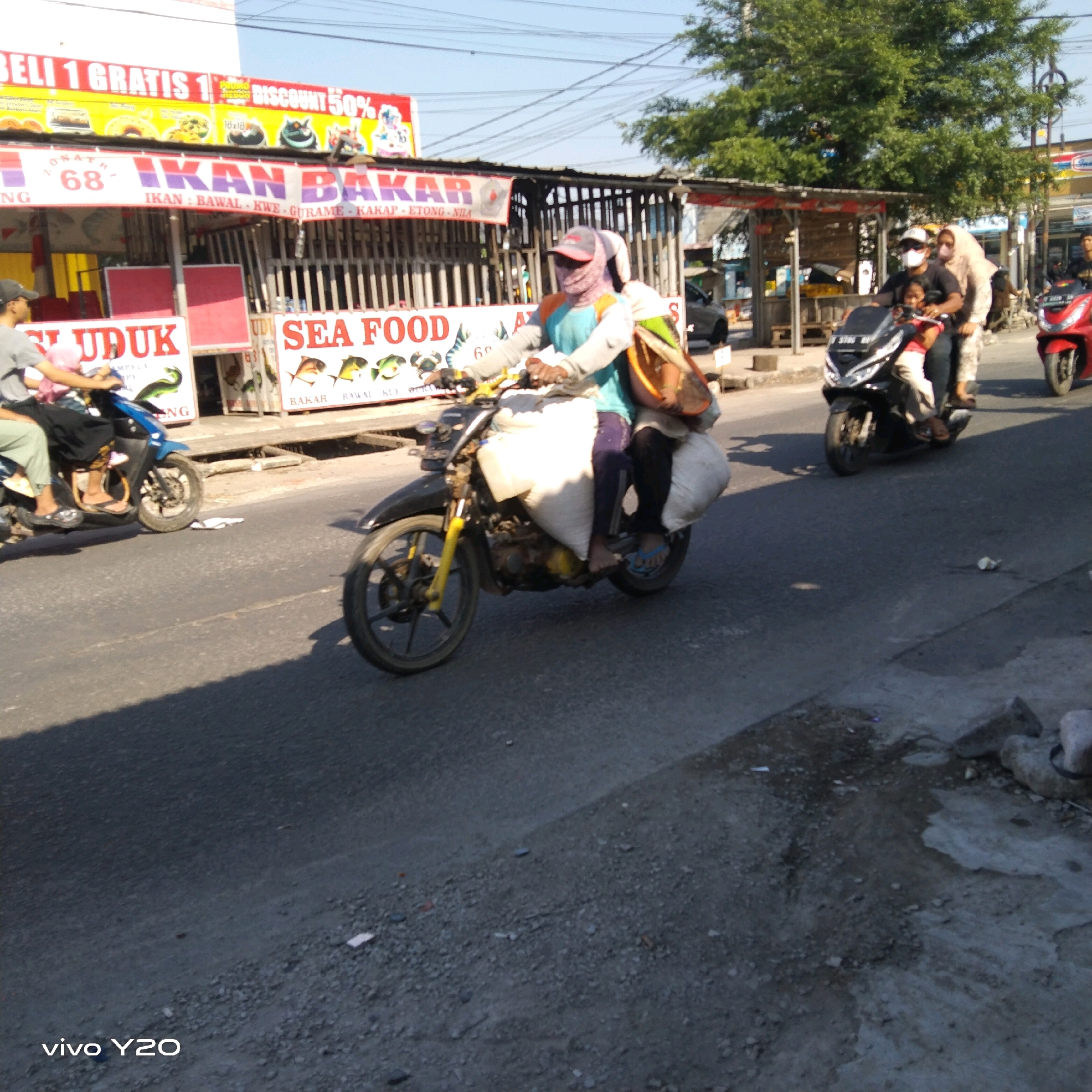 In an outdoor, urban environment under a clear sky, a person is riding a motorcycle on a paved road. The rider is wearing a light blue long-sleeved shirt, dark pants, and a red and grey cap. They are also wearing a face covering. The motorcycle is dark-colored with yellow accents on the front fork. The rider is carrying two large white sacks, one on each side of the motorcycle, and a brown bag on their back. In the background, there are several street stalls with signs advertising "IKAN BAKAR" (grilled fish) and "SEA FOOD". One stall has a sign that says "BELI 1 GRATIS 1 DISCOUNT 50%". There are other motorcycles and people in the background, some of whom are also on motorcycles. The road surface is asphalt with some gravel and debris. The scene appears to be a street in a market area.