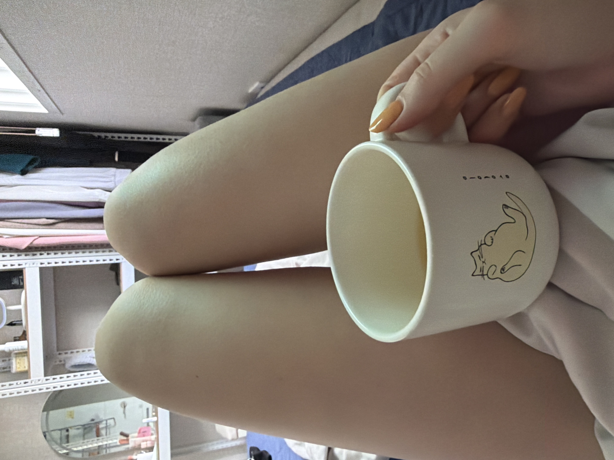 A person's legs are visible in the foreground, with a white mug of yellow liquid, possibly tea, resting between them. The mug has a drawing of a cat on it and some Korean text. The person's hand, with orange nail polish, is holding the mug. In the background, a shelf unit with various items and clothing hanging on a rack is visible, along with a window. The scene appears to be indoors, in a residential setting in Dongducheon-si, South Korea.